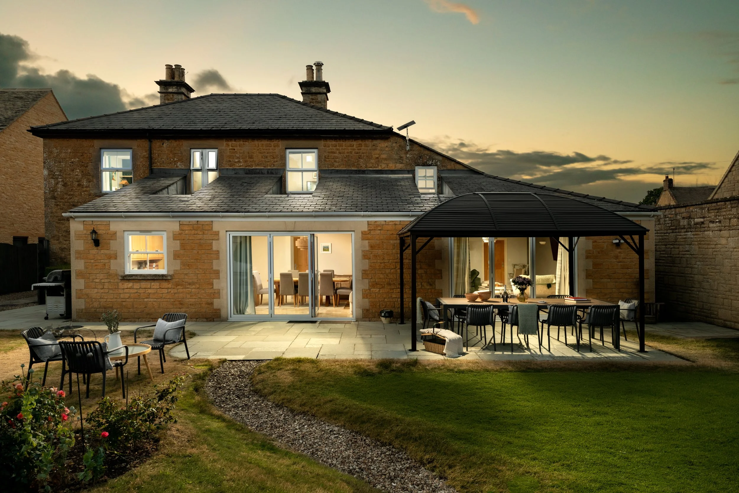Backyard patio area with outdoor dining table and chairs, garden plants, and a view of the back of a brick house with large windows and a covered patio at sunset.