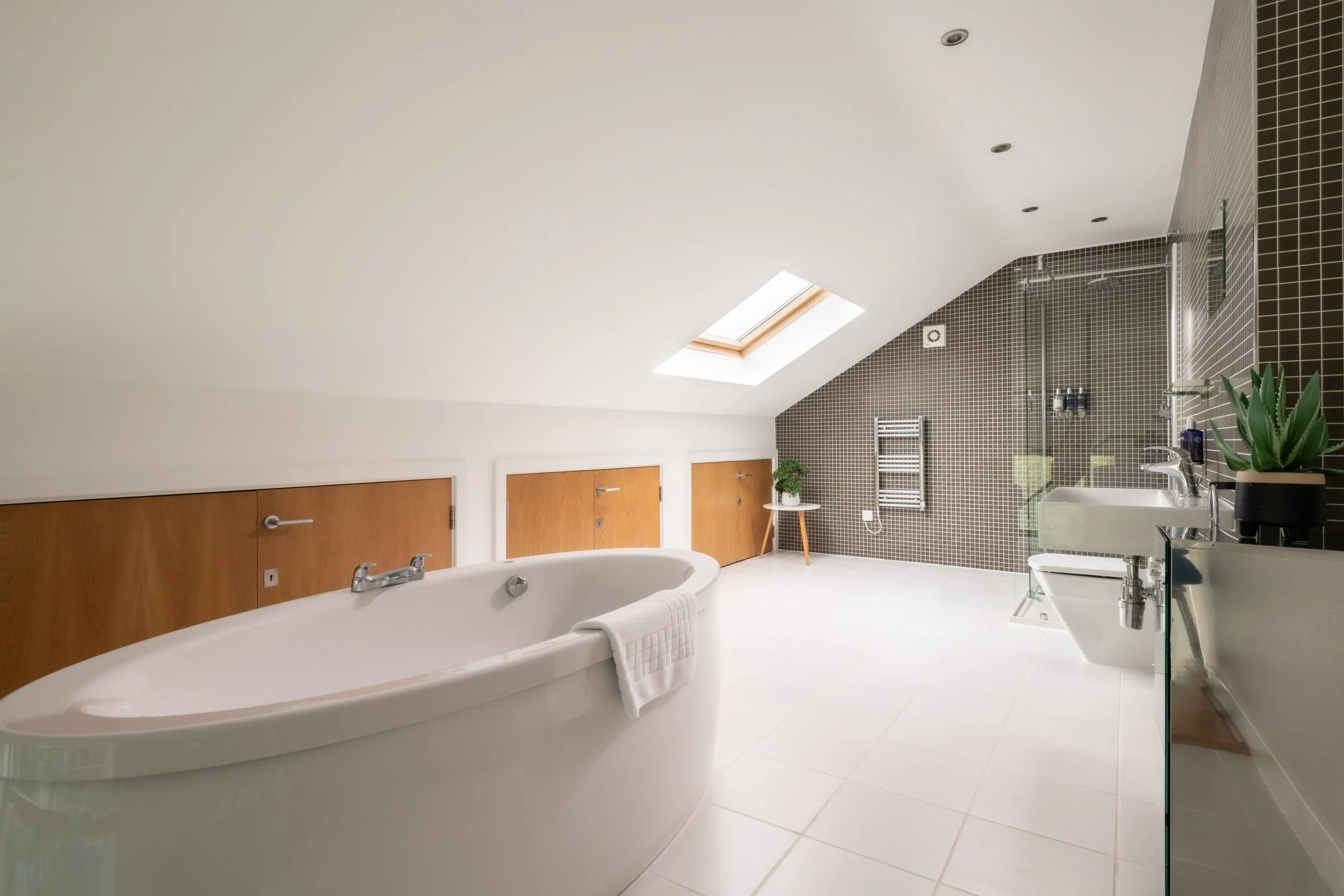 Modern bathroom with a white bathtub, wooden doors, a skylight, black tiled wall with a shower area, potted succulent plant, and white floor tiles.