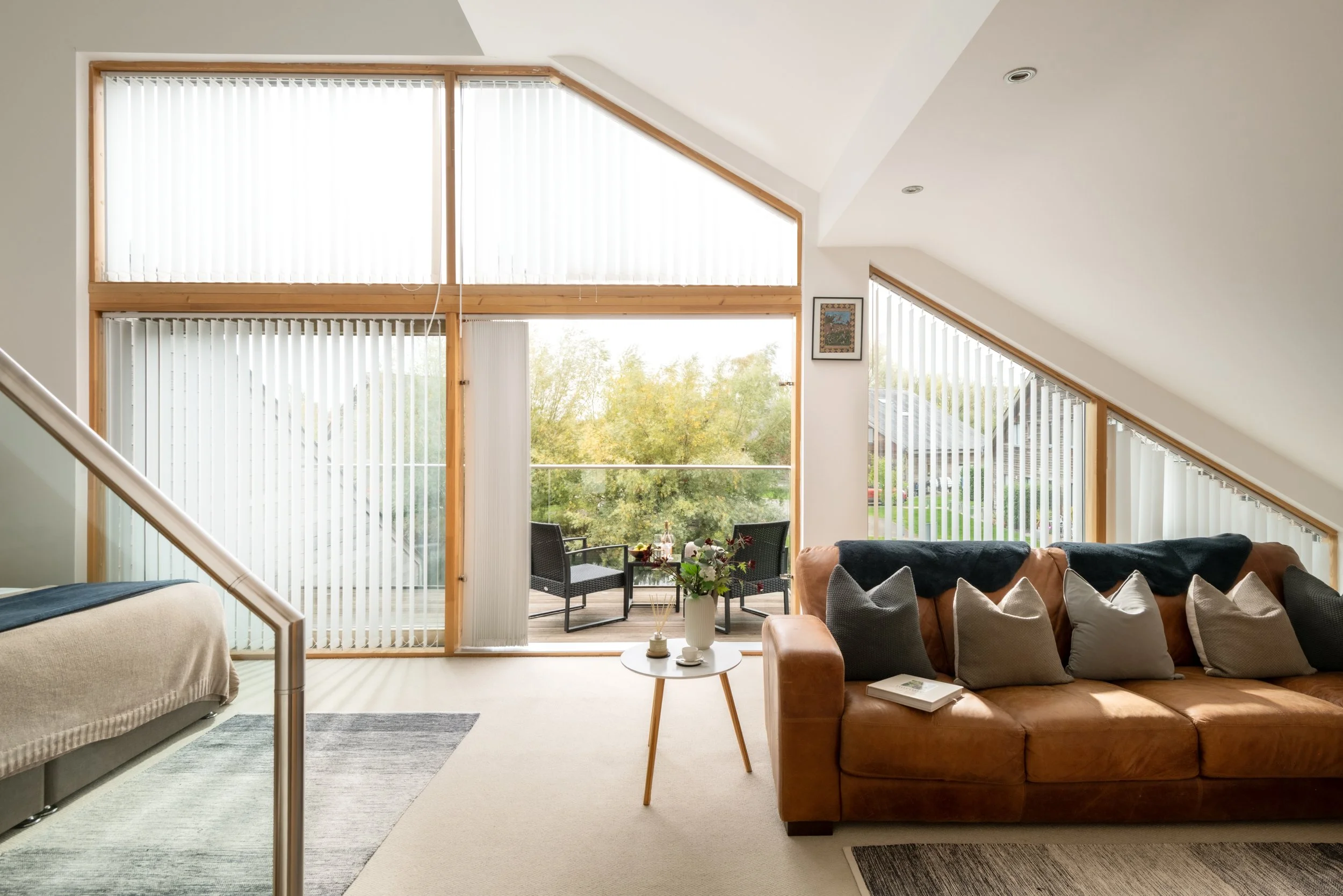 Living room with large windows, a brown leather sofa with pillows, a beige armchair, a small round white coffee table with a vase of flowers, a living room with a view of trees outside, vertical blinds on the windows, and a piece of framed artwork on