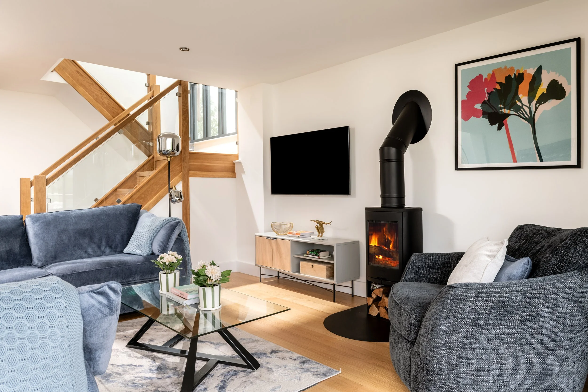 A cozy living room with blue velvet sofa, black armchair, glass coffee table with plants, and modern decor. A wood stove with a fire, framed artwork, and staircase with glass railing are visible.