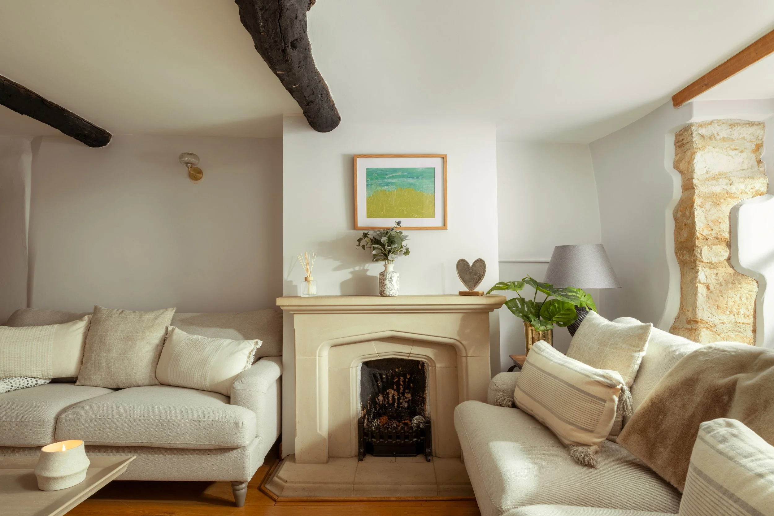 A cozy living room with two cream-colored sofas with cushions, a fireplace with a mantel decorated with a potted plant and a heart-shaped ornament, a painting of a green landscape hanging above the mantel, a gray lampshade beside the fireplace, a sma