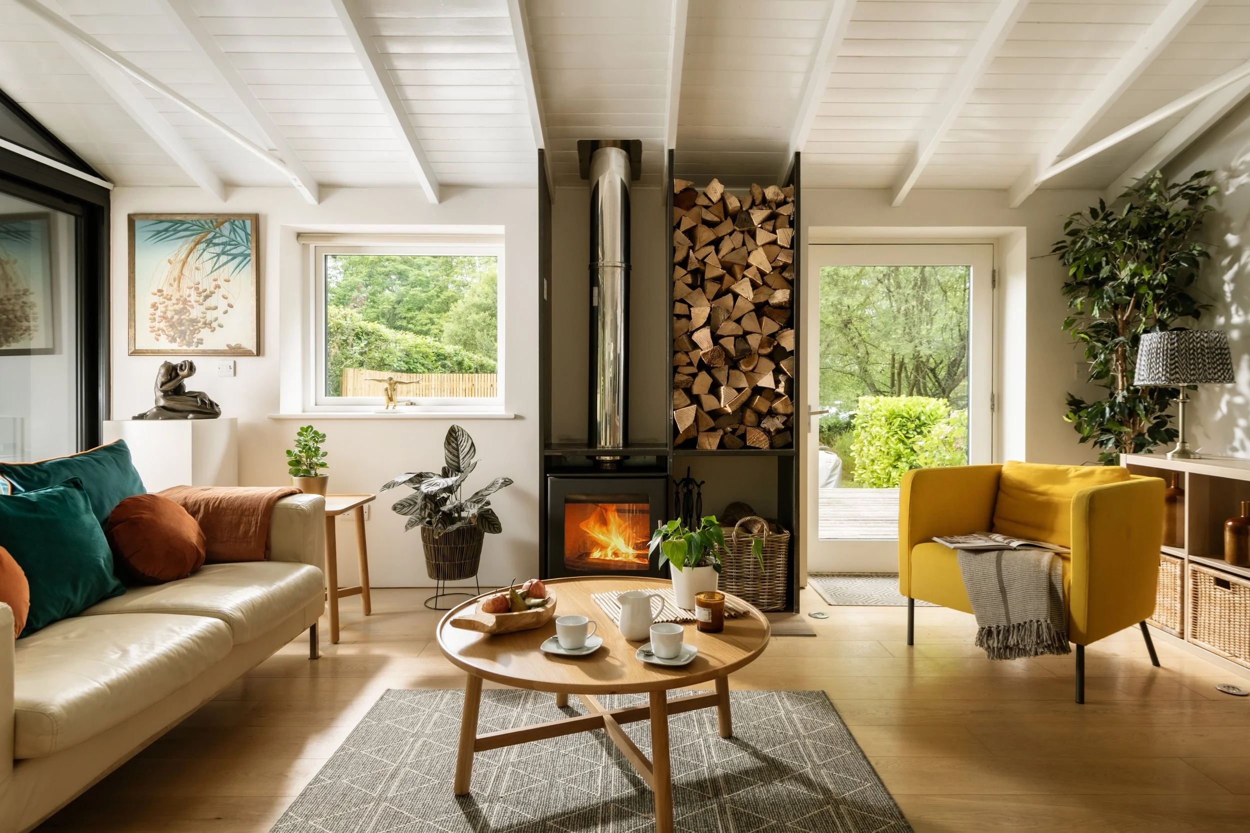 A cozy living room with a white couch, colorful pillows, a wooden coffee table with cups and a snack, a fireplace, wood storage, and large windows with greenery outside.