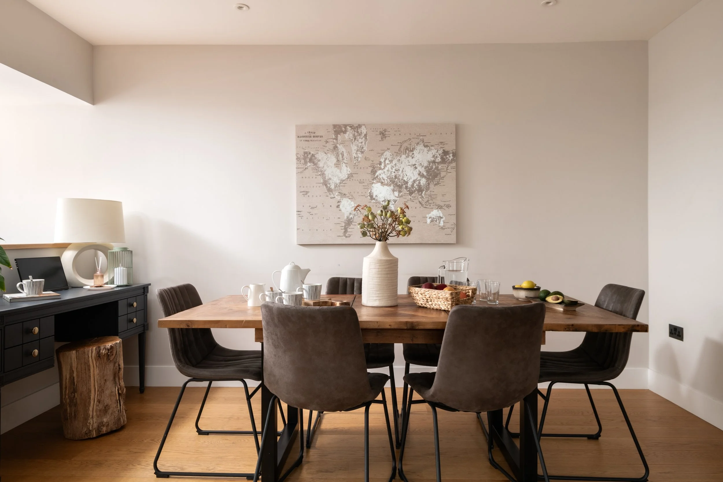 A dining room with a wooden table, six gray velvet chairs, a black sideboard with gold knobs, and a white vase with a floral arrangement. A world map hangs on the white wall behind the table, and the room is well-lit with natural light.