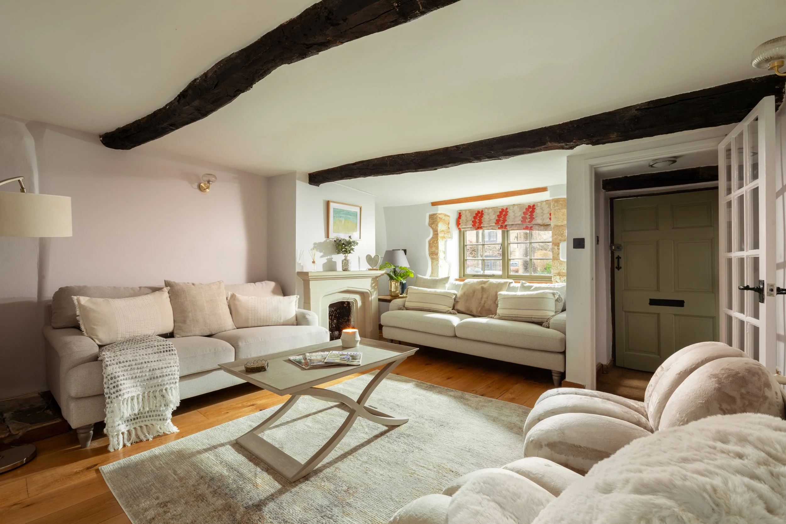 A cozy living room with exposed dark wooden beams on the ceiling, cream-colored sofas, a coffee table, a fireplace, a window with floral curtains, and various decorative items including framed artwork and plants.