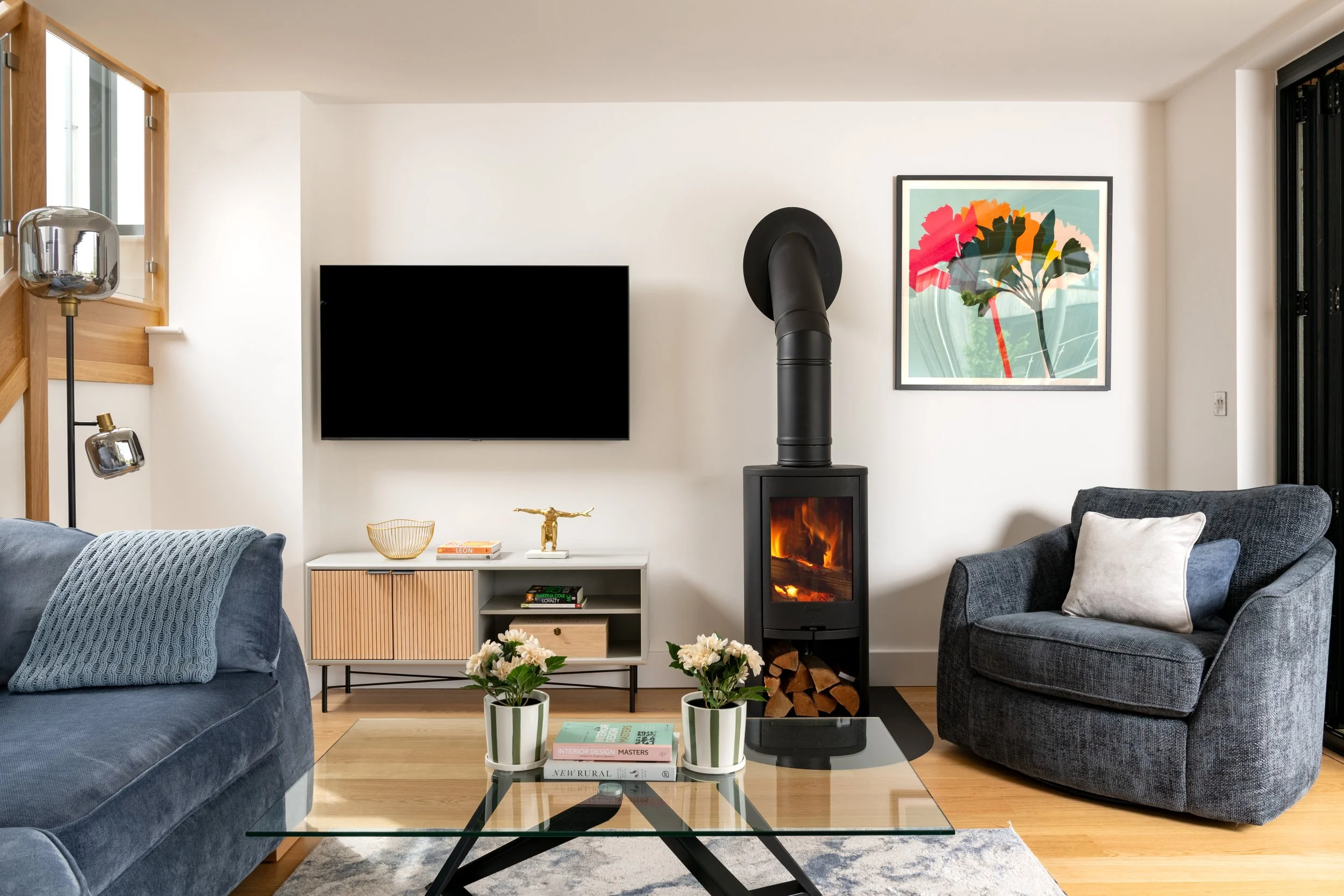 Living room with a black wood stove, framed artwork, flat-screen TV, gray armchair, blue sofa with knitted blanket, plants, books, and decorative objects.