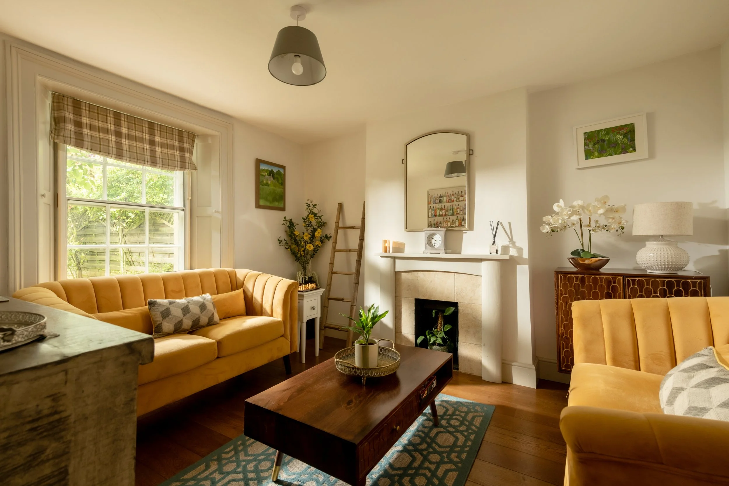 Living room with a large window, yellow sofas, a coffee table, potted plants, a ladder, framed artwork, and a fireplace with a mirror above it, decorated with flowers and a lamp.