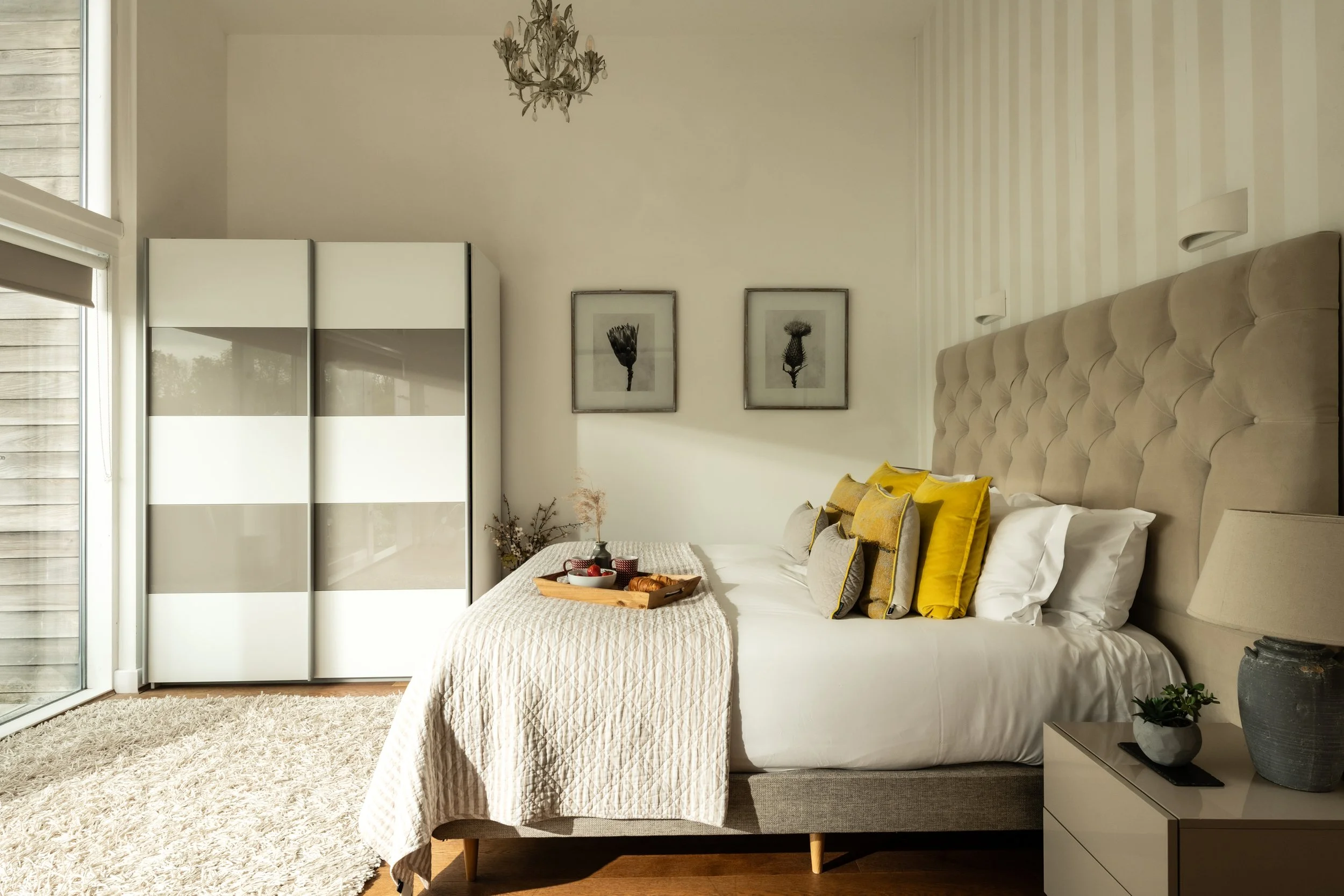 A bedroom with a large bed featuring a tufted beige headboard, yellow and beige pillows, and a cream quilt. On the bed, there is a wooden tray with breakfast items. To the right of the bed, there is a nightstand with a gray vase, a small plant, and a