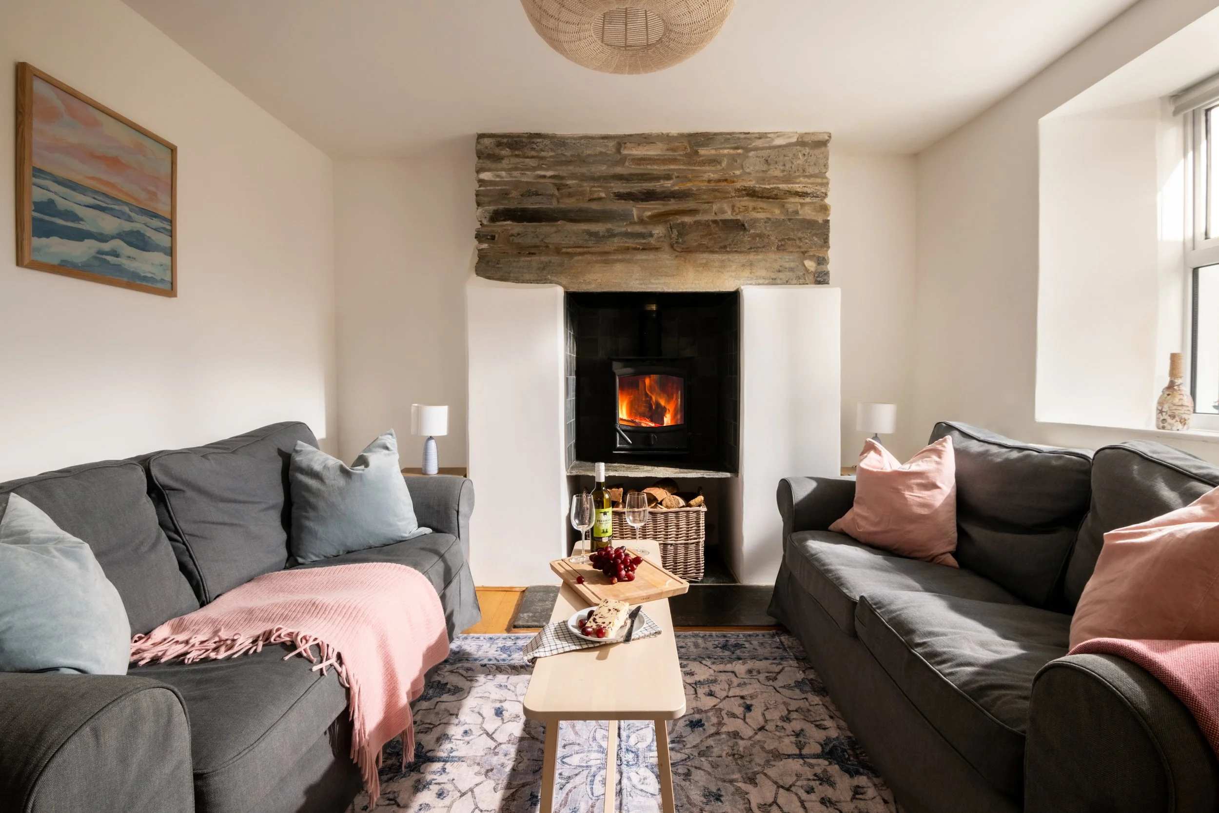 Living room with a fireplace, two gray sofas with cushions, a coffee table with wine and grapes, a painting of a seascape on the wall, and daylight coming through a window.