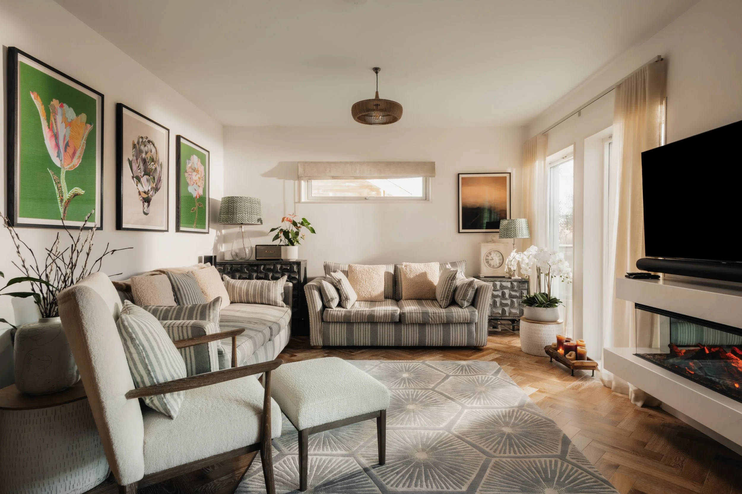 Cozy living room with striped sofas, artwork on white walls, a fireplace with candles, potted plants, and large windows with curtains.