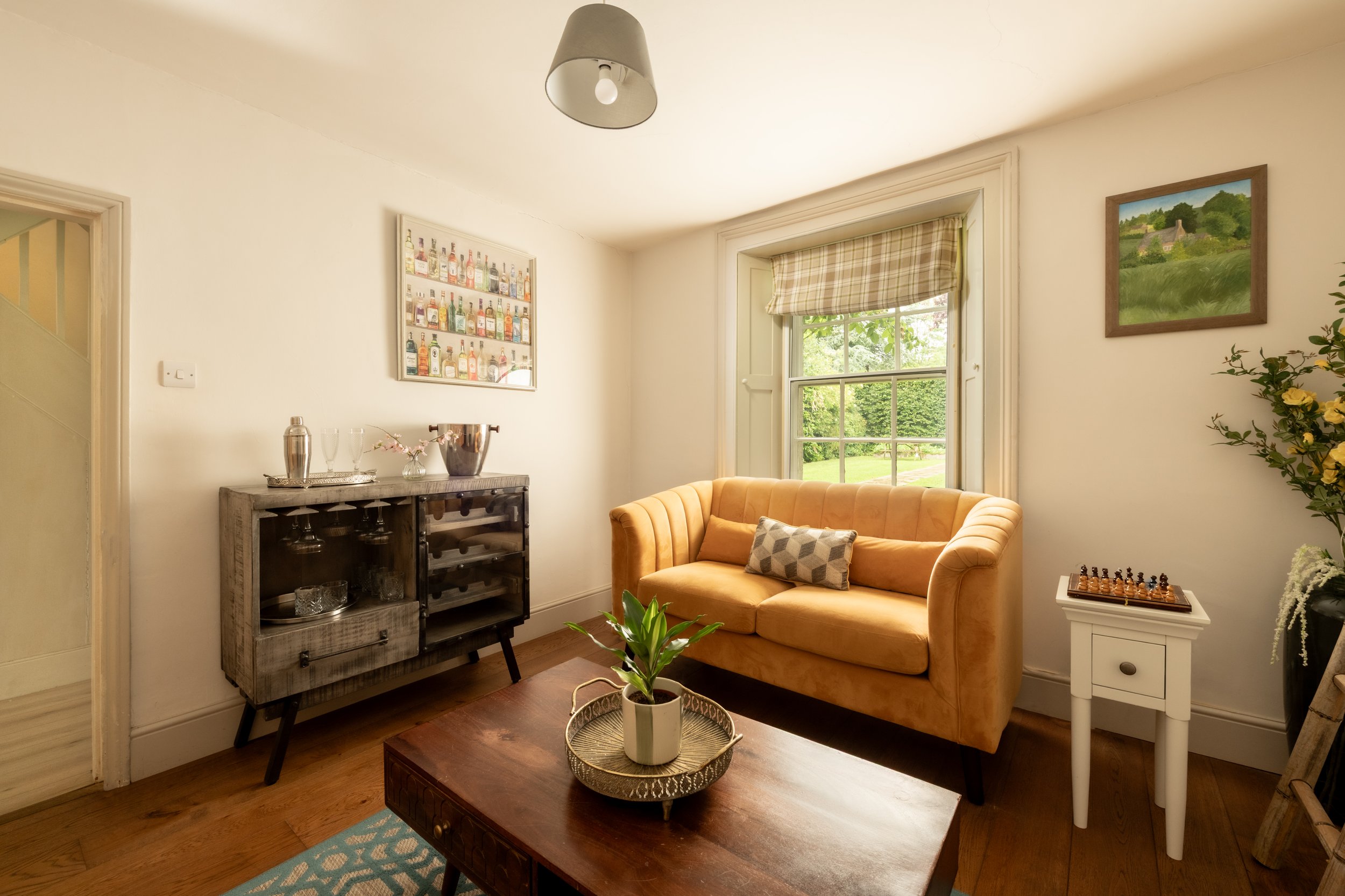 Living room with a yellow sofa, a wooden coffee table with a potted plant, a small white side table, a wooden cabinet with glasses and bottles, framed artwork on the walls, a window with plaid curtains, and a chessboard on the side table.