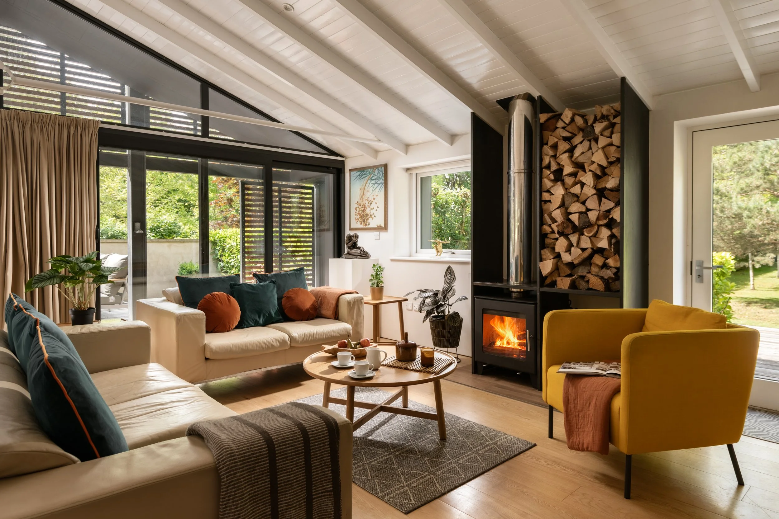 Living room with beige sofas, colorful throw pillows, a round coffee table with cups and snacks, a yellow armchair, a wood-burning fireplace with stacked firewood, large windows with views of green trees, framed artwork on the wall, and potted plants