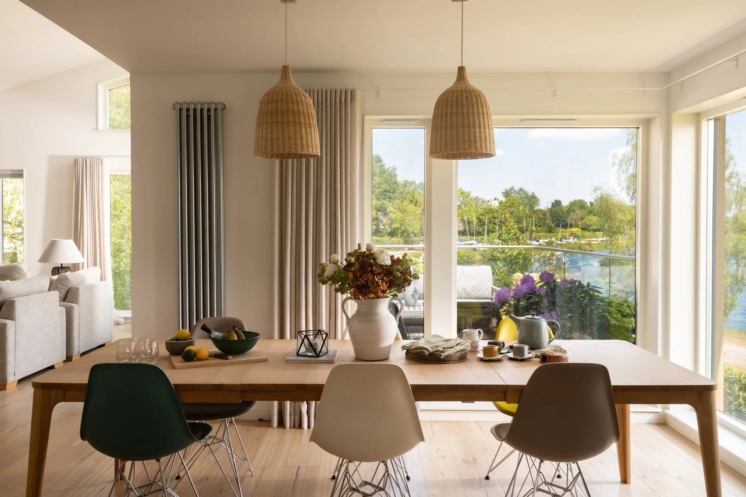 A modern, bright living room with large windows overlooking greenery and a water scene, featuring beige sofas, curtains, wicker pendant lights, and a dining table with various decorative items and fruits.