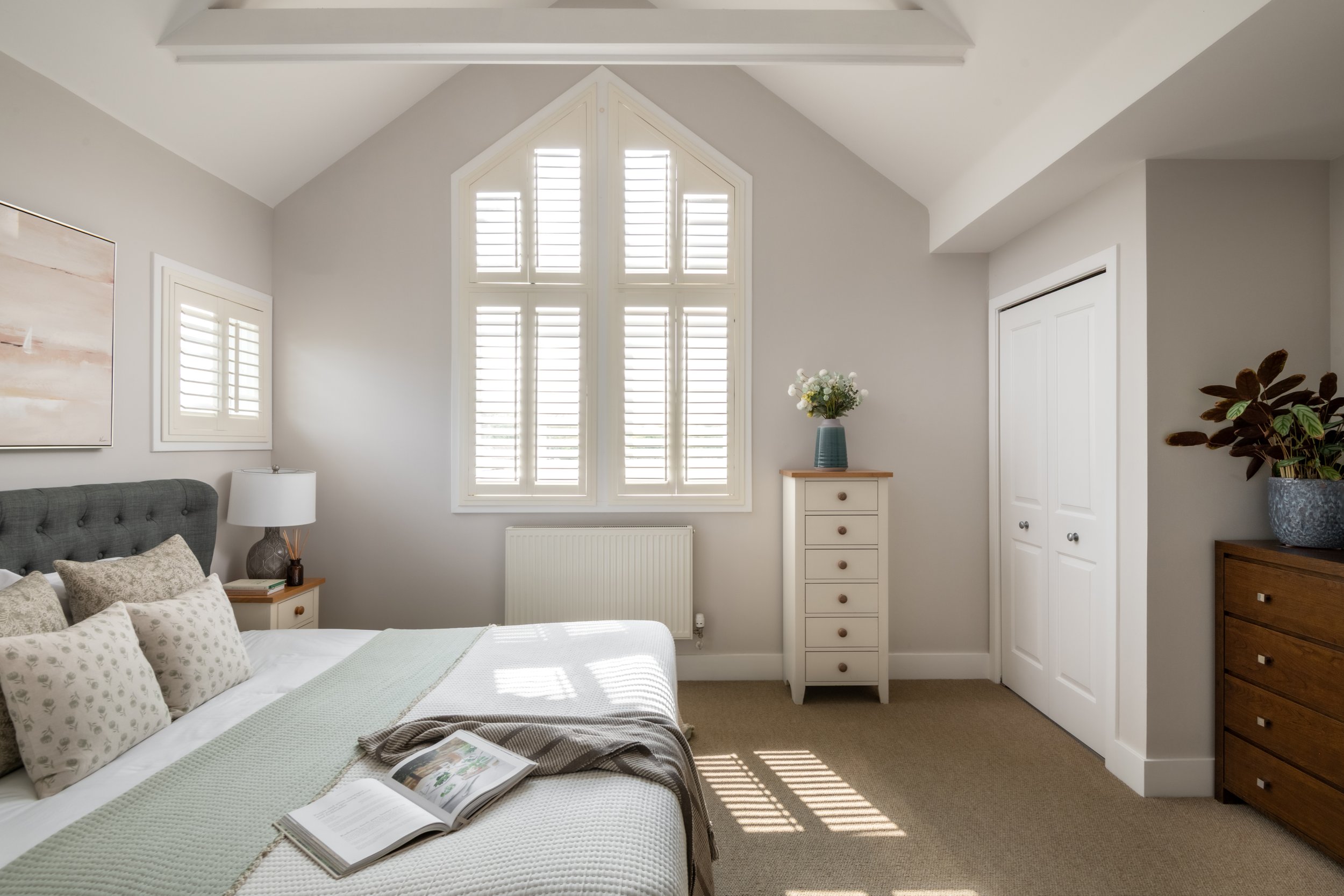 A bedroom with a bed on the left, sunlit through large window with white shutters, a white dresser with flowers, and a closet with double doors, beige carpet, and neutral-colored walls.