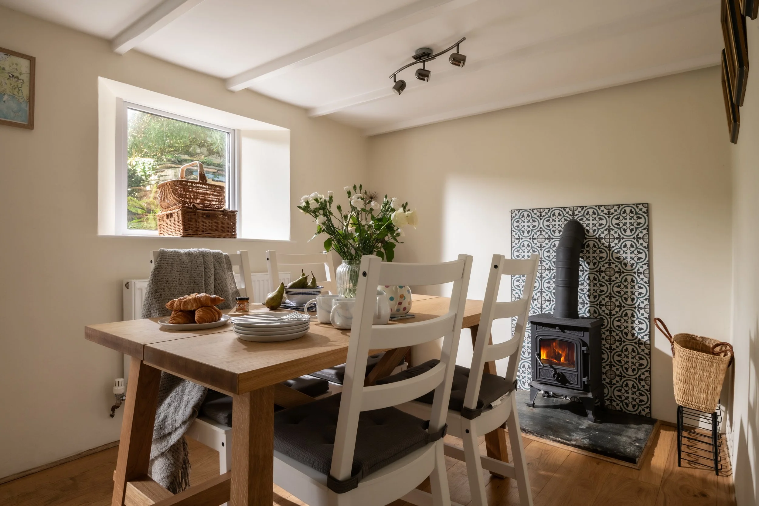 Cozy dining room with a wooden table set with plates, mugs, and croissants, white chairs, a window with a woven basket on the sill, a vase with white flowers, and a wood stove with a patterned tile back, a basket, and a wall art. sunlight streams in 