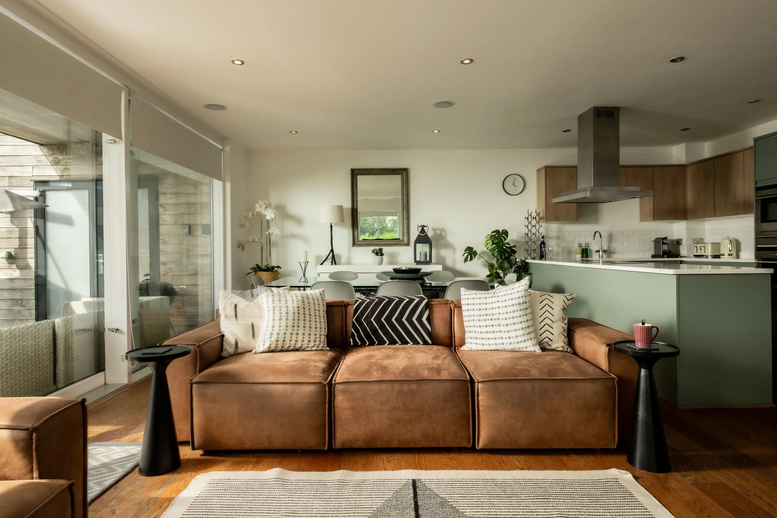 Modern living room with brown sofa, decorative pillows, black side tables, and a view into the kitchen area with wooden cabinets, a stainless steel range hood, and various small appliances.