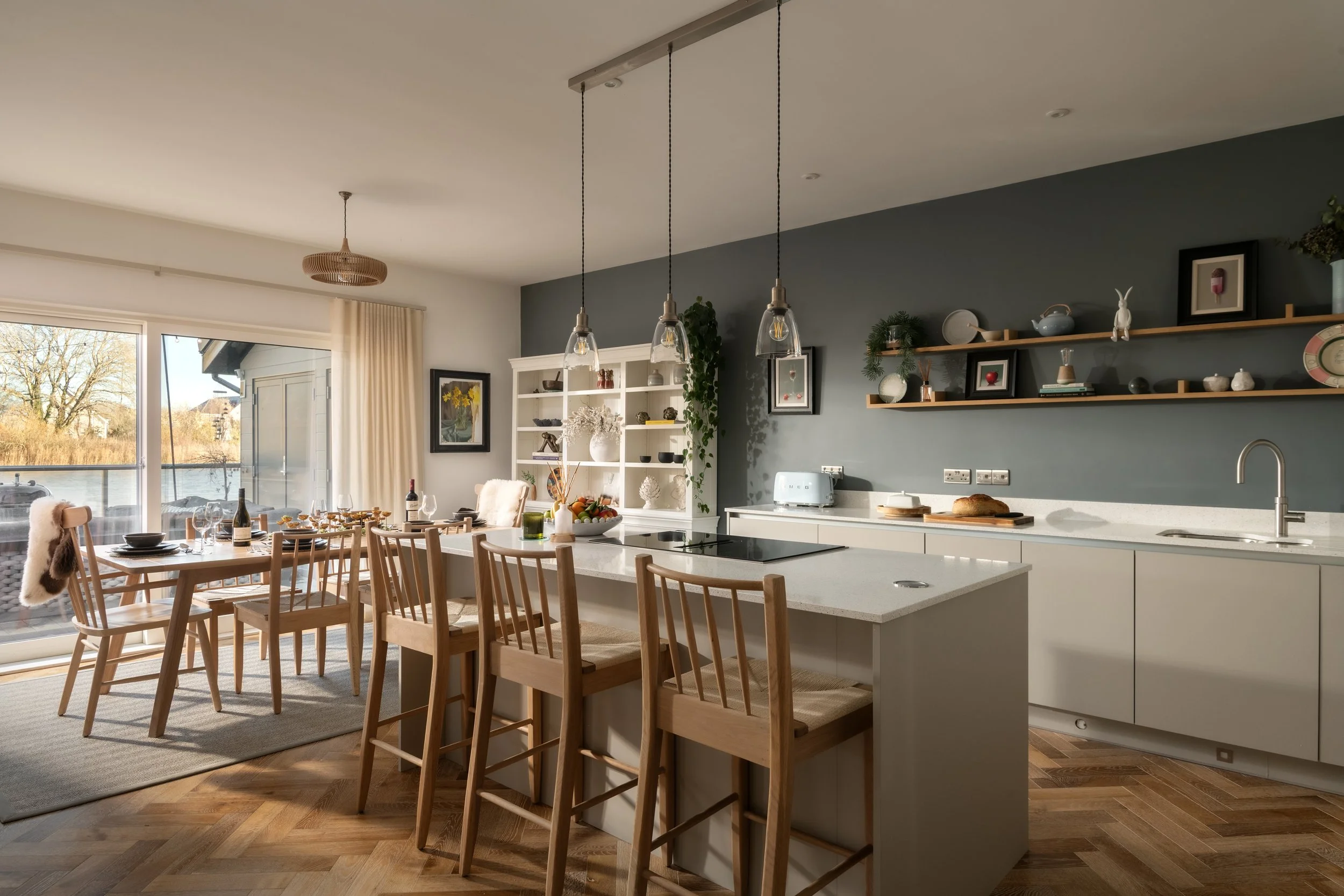 A bright, modern kitchen and dining area with large windows revealing an outdoor view, wooden chairs around a dining table set with wine glasses and food, white cabinetry, shelves with decorative items, and pendant lights hanging above the kitchen is