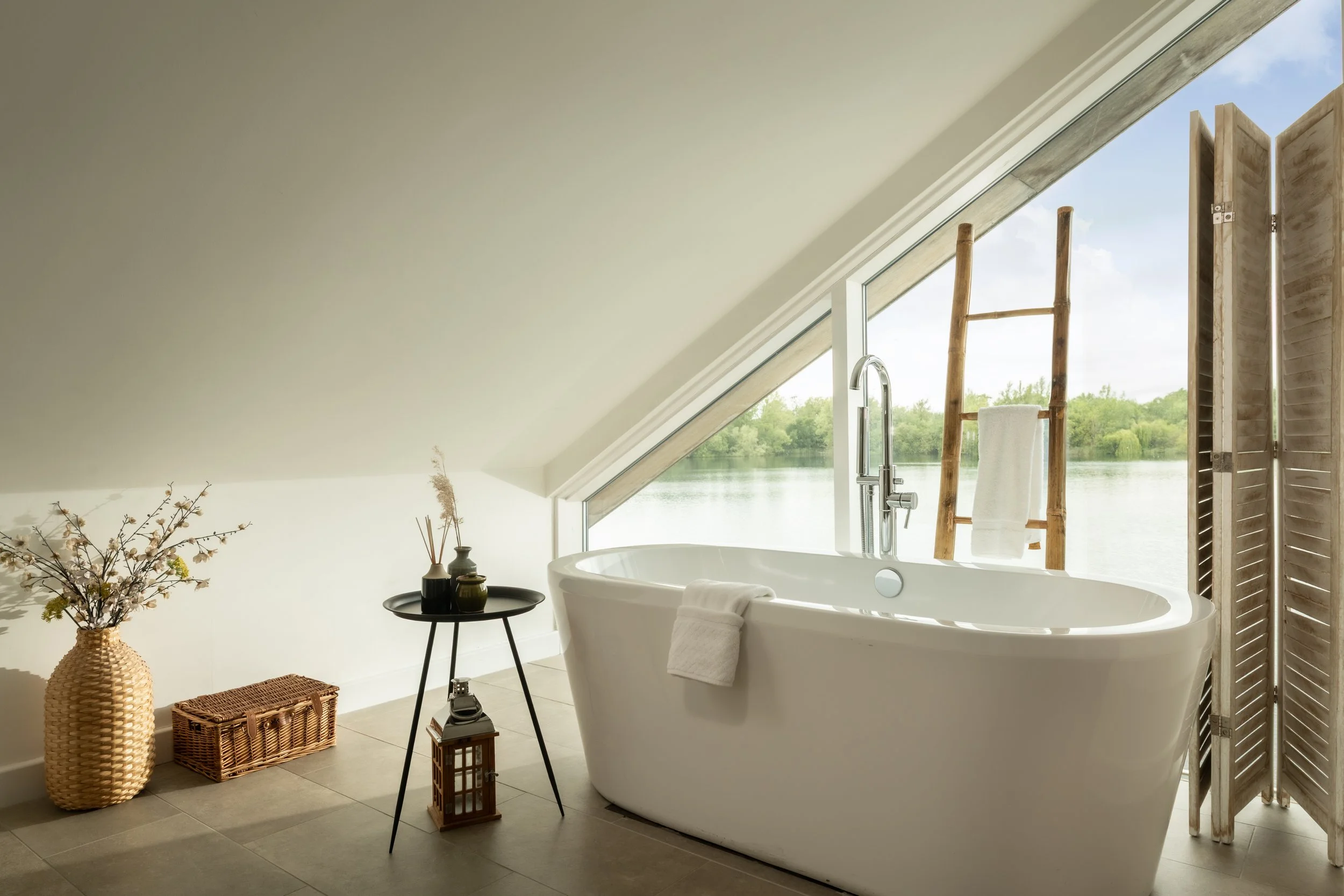 A modern bathroom with a large freestanding bathtub positioned near a sloped ceiling window overlooking a lake and trees. Decor includes a vase with branches, a wicker basket, a small black side table, and a wooden lantern. A wooden ladder with a tow