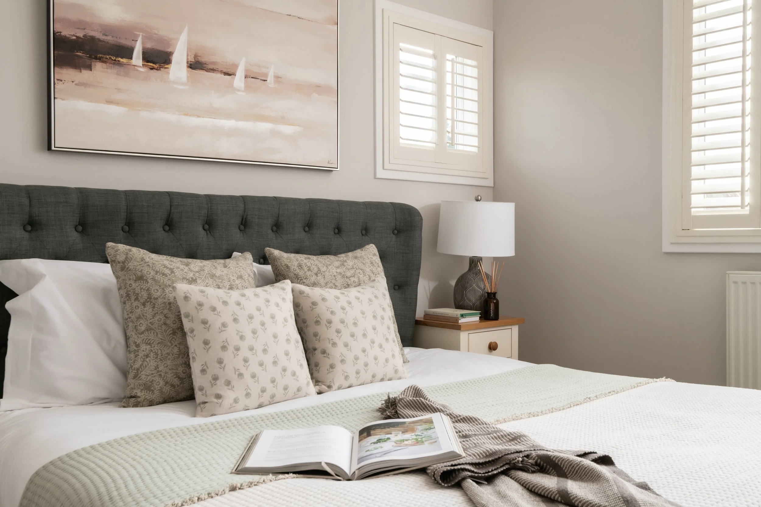 A neatly made bed with a tufted gray headboard, decorated with beige and white patterned throw pillows, a white blanket, and an open book on top. Next to the bed is a small white nightstand with a dark vase, a stack of books, and a patterned table la