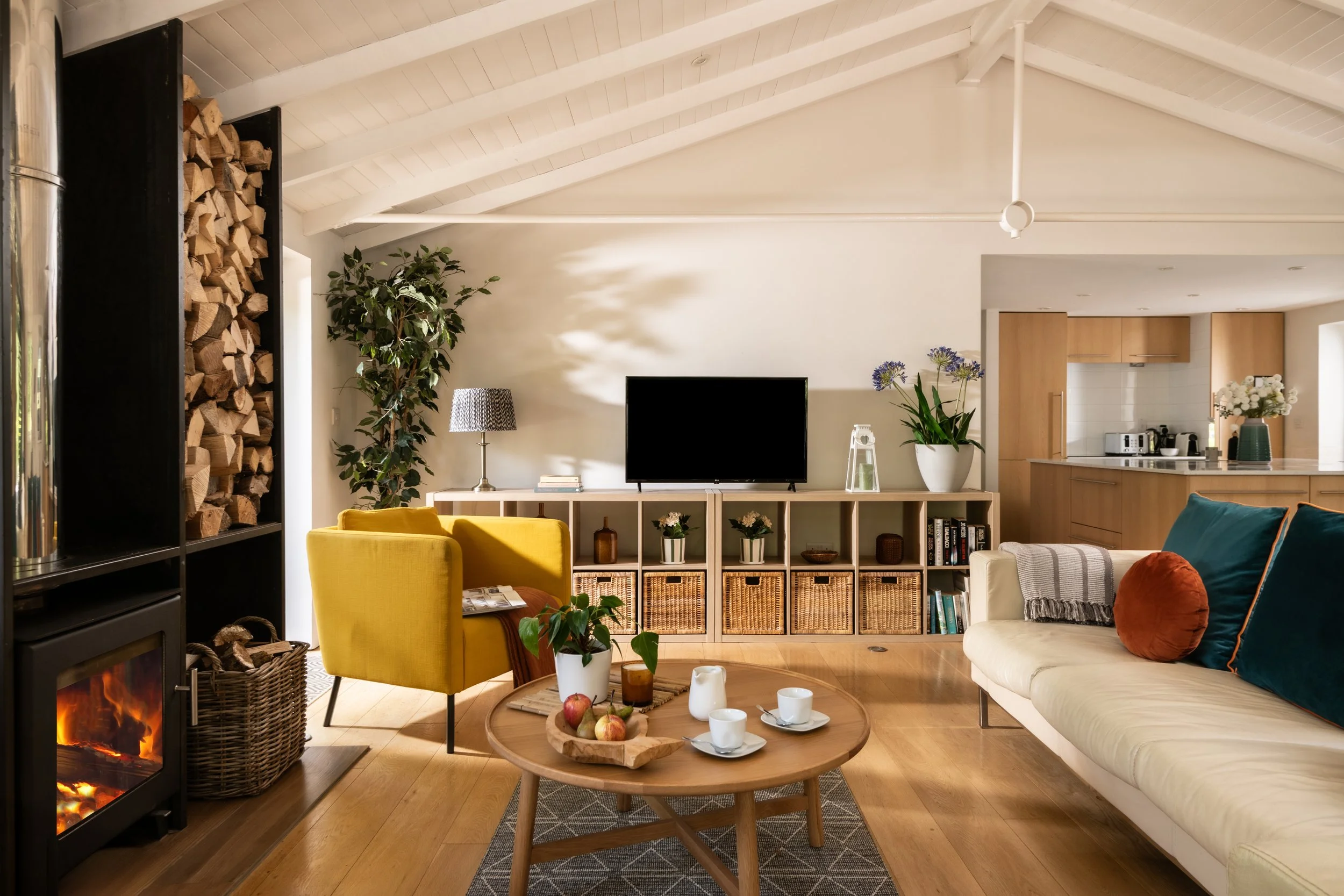 Living room with a wood-burning fireplace, yellow armchair, sofa with colorful pillows, coffee table with apple and cups, TV on a cabinet, bookshelf, and kitchen in the background.