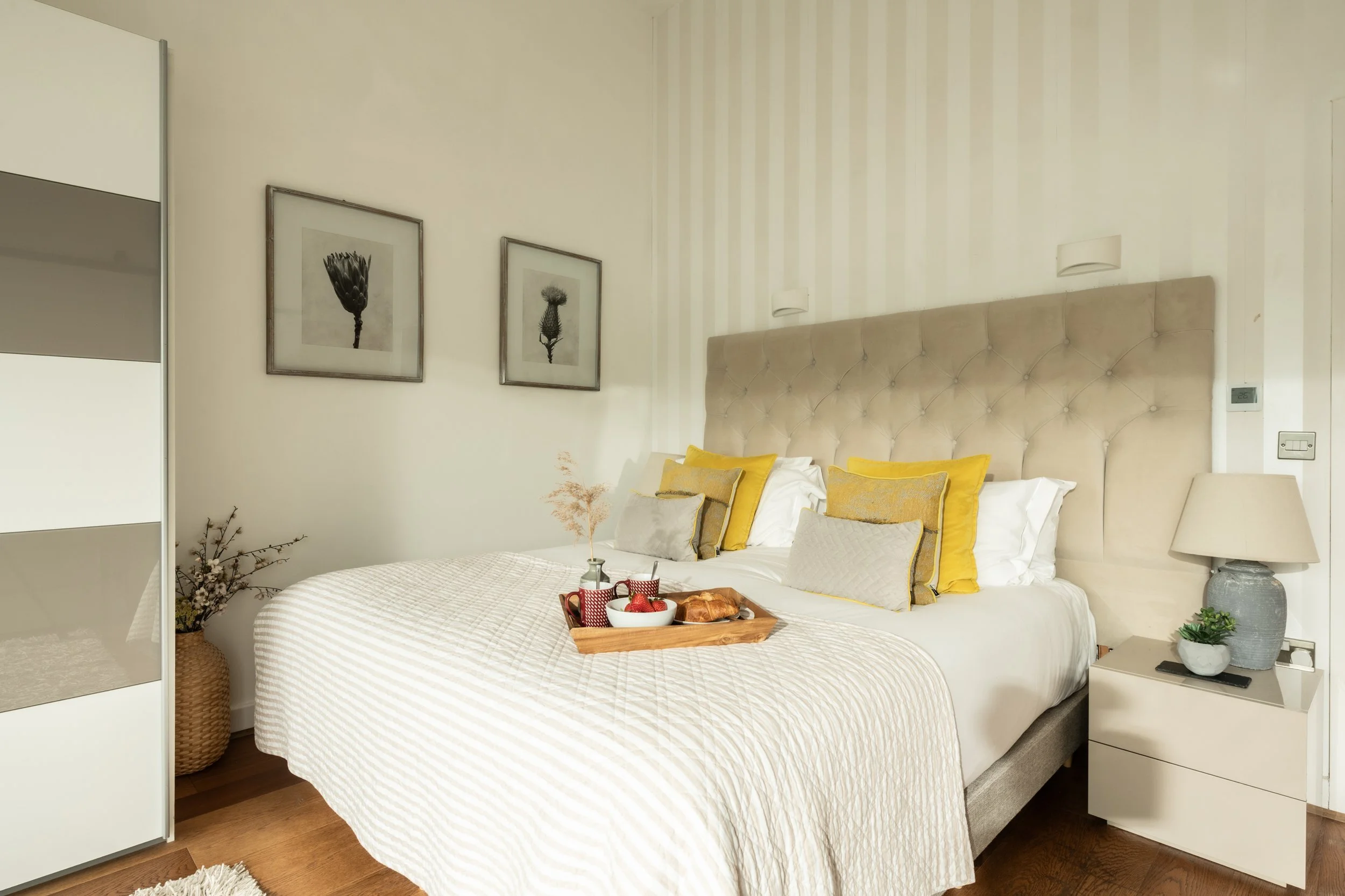 A cozy bedroom with a large bed, beige upholstered headboard, yellow and beige accent pillows, white bedding, a bedside table with a lamp, framed botanical pictures on the wall, and a breakfast tray with strawberries and pastries.