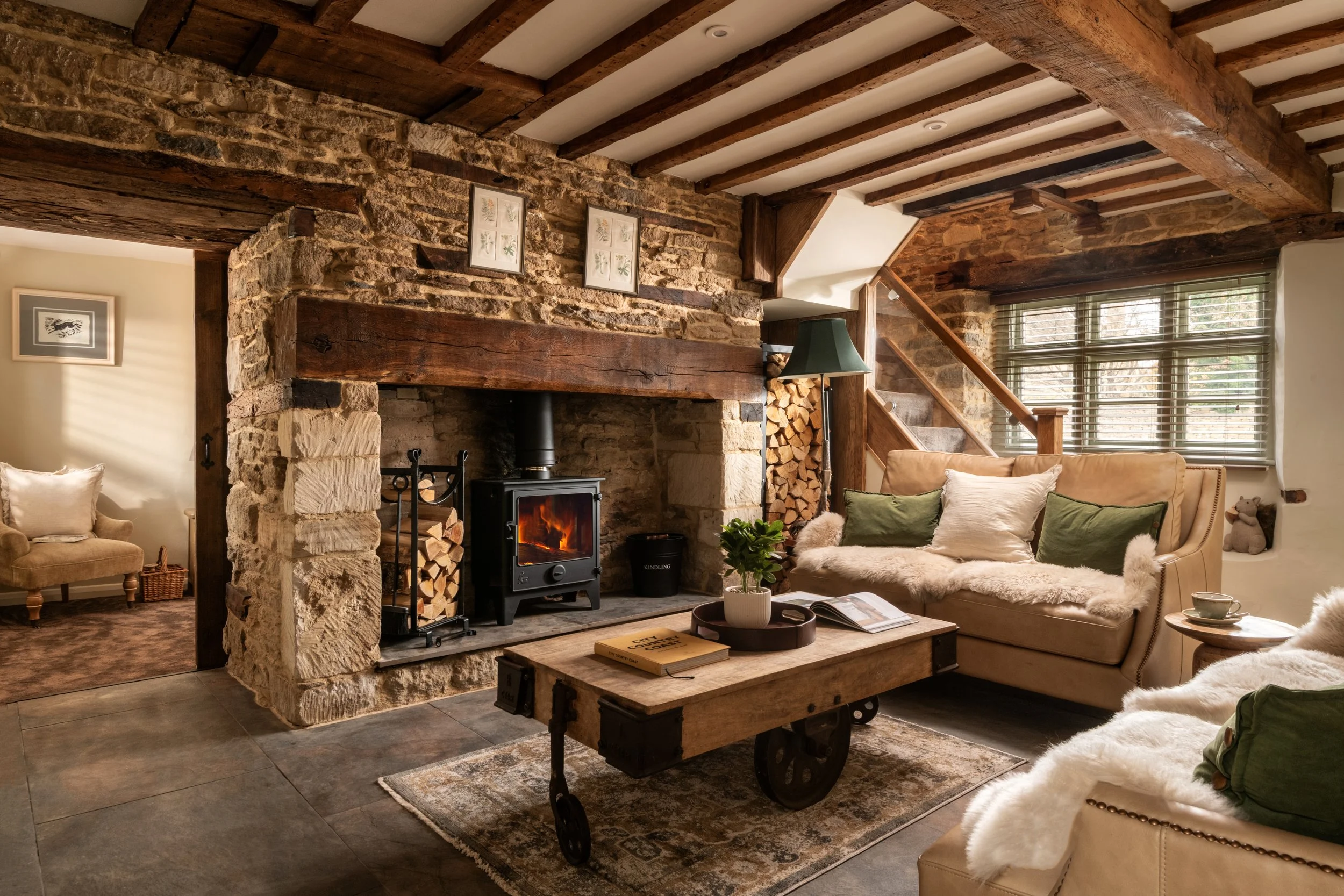 Cozy living room with exposed wooden beams, stone fireplace, wood logs, wood stove, plush beige sofa with pillows, coffee table with books and a plant, window with blinds, and decorative framed art on the walls.