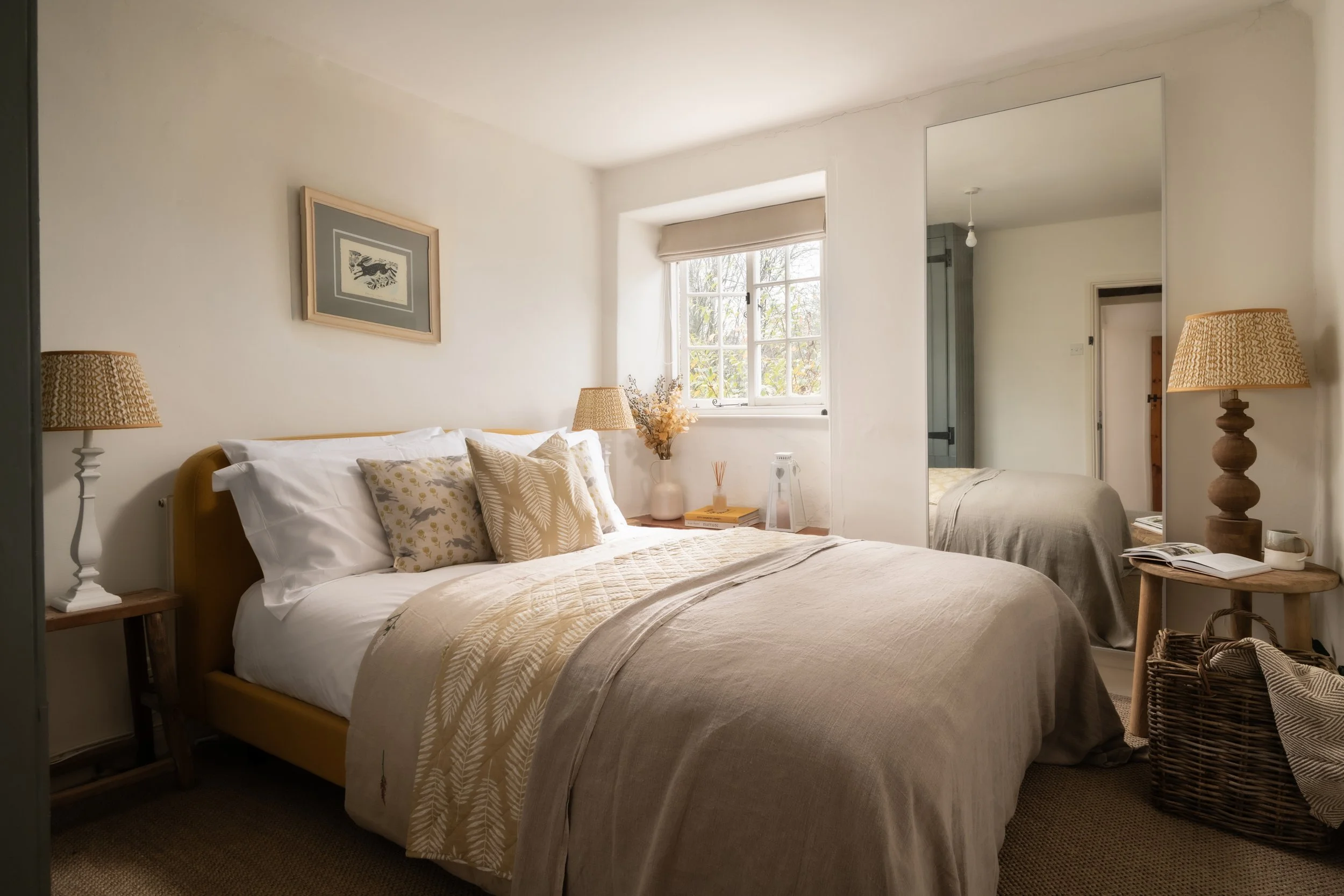 A cozy bedroom with a large bed covered in beige and white bedding, decorative pillows, and a wooden headboard. There are two bedside tables with matching lamps and a framed picture on the wall above the bed. A window with a beige shade lets in natur