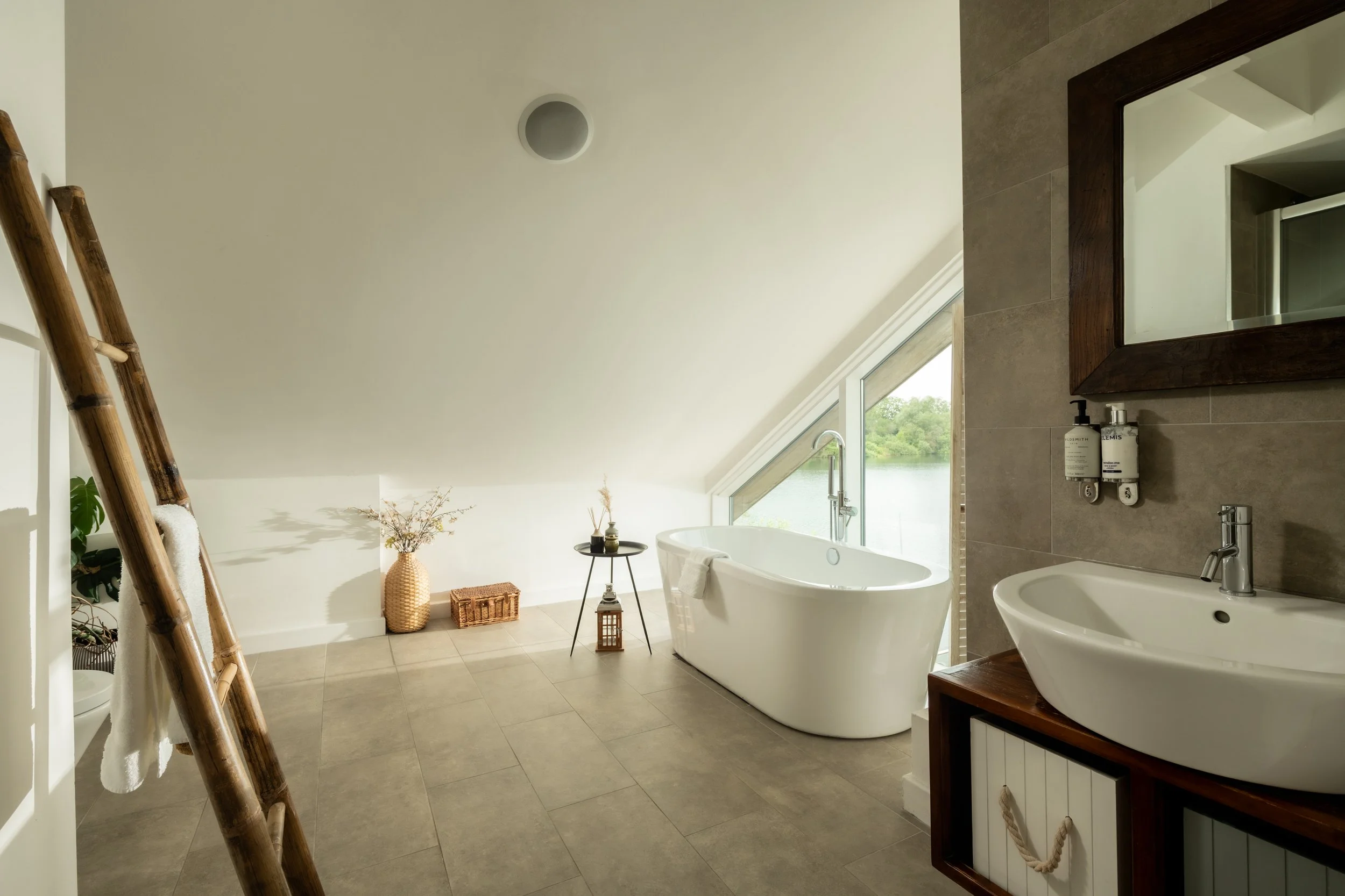 A modern bathroom with a freestanding bathtub, a sink with a wooden cabinet, a large mirror, and decor including vases, a lantern, and a small table near a large window with a view of greenery outside. There is a wooden ladder with towels on the left