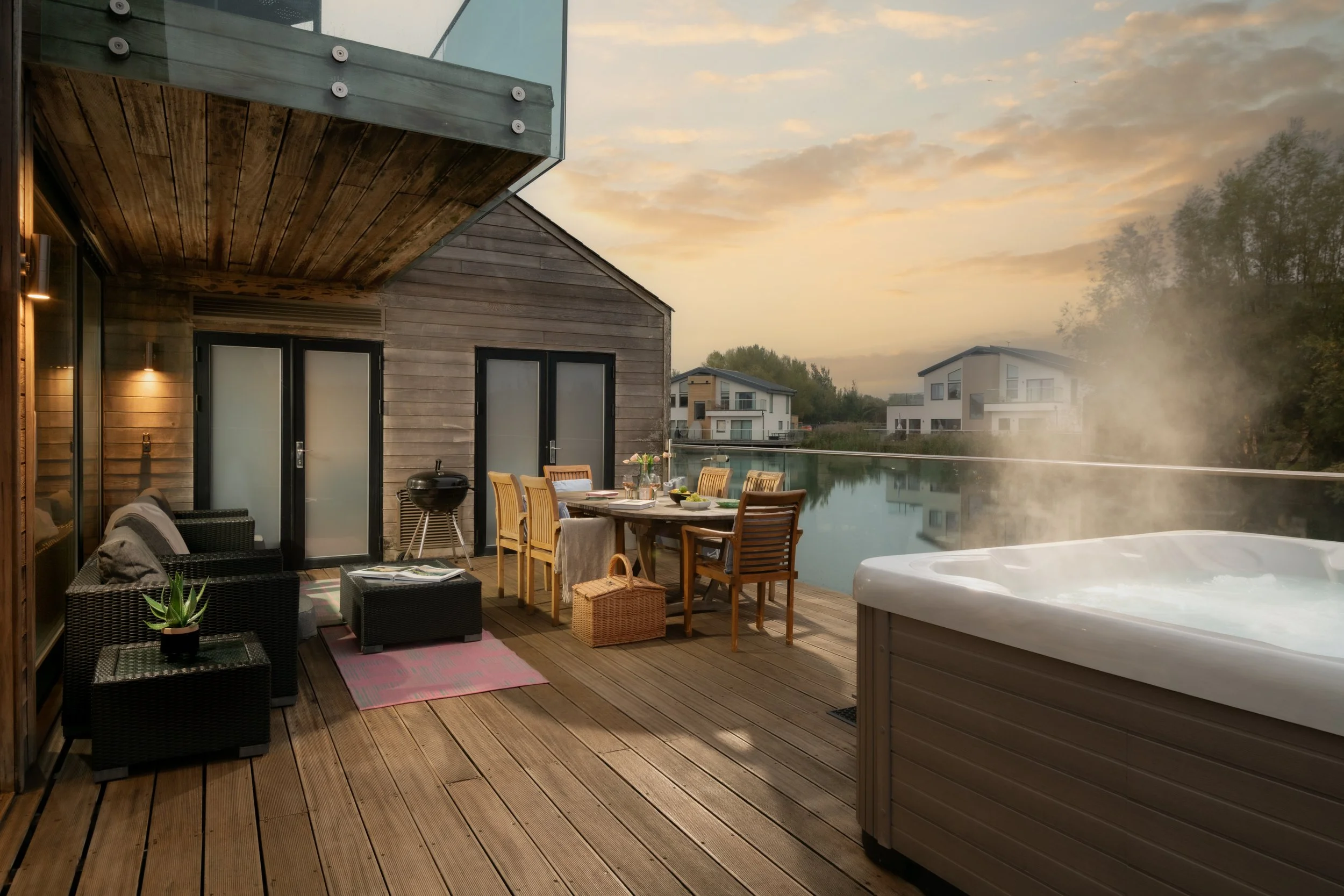 A modern outdoor balcony at sunset with a wooden deck, outdoor furniture, a hot tub, and a glass railing, overlooking neighboring houses and trees.