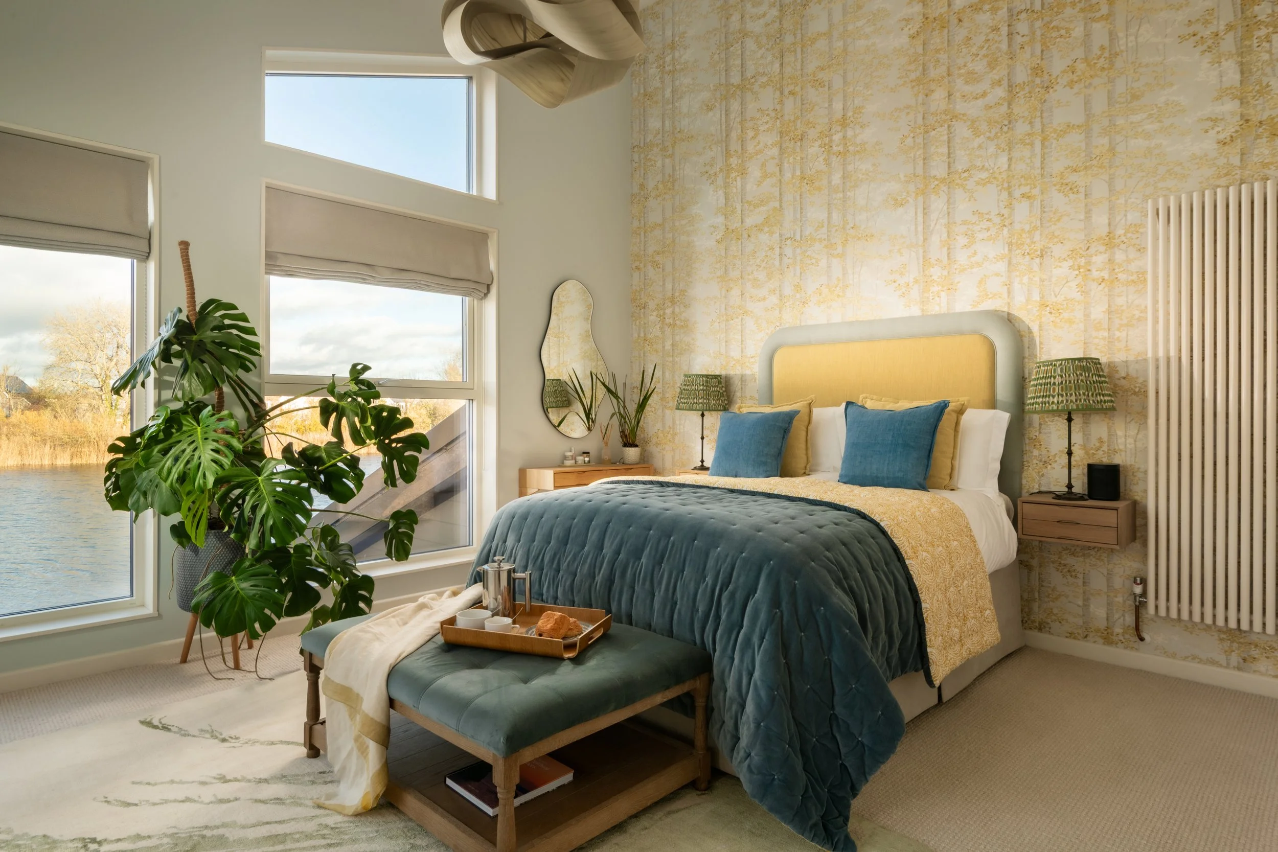 Bedroom with large windows overlooking a body of water, with a bed covered in a blue quilt and yellow and blue pillows, a wooden side table with a tray of breakfast, a large plant, and a mirror on the wall.