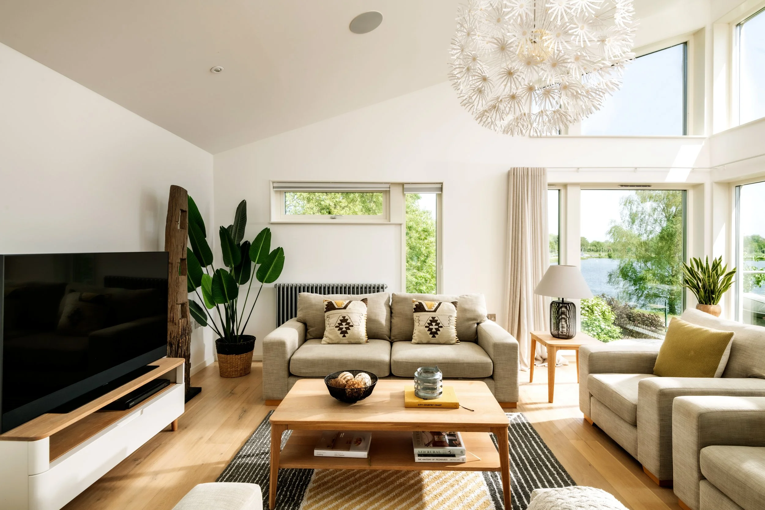 Living room with beige sofa, large windows overlooking greenery and water, white walls, wooden coffee table, television, green indoor plants, modern chandelier, patterned throw pillows, natural light.