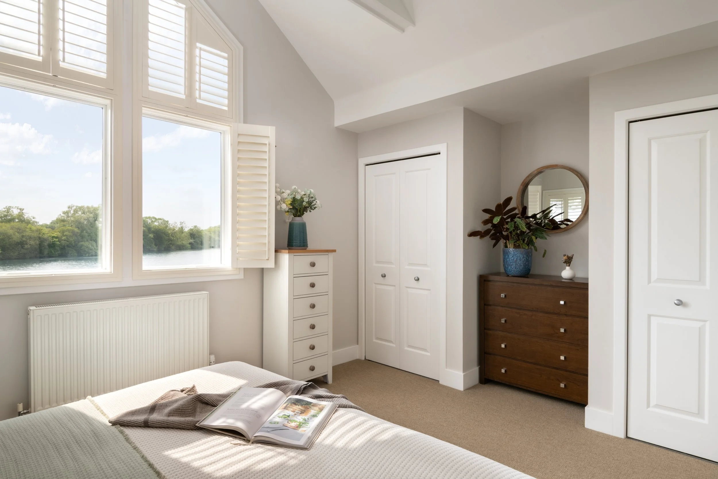 A cozy bedroom with large windows that have white shutters, showing a view of a river and trees outside. Inside, there is a bed with an open magazine, a white chest of drawers with a blue vase of flowers, a wooden dresser with a potted plant and mirr