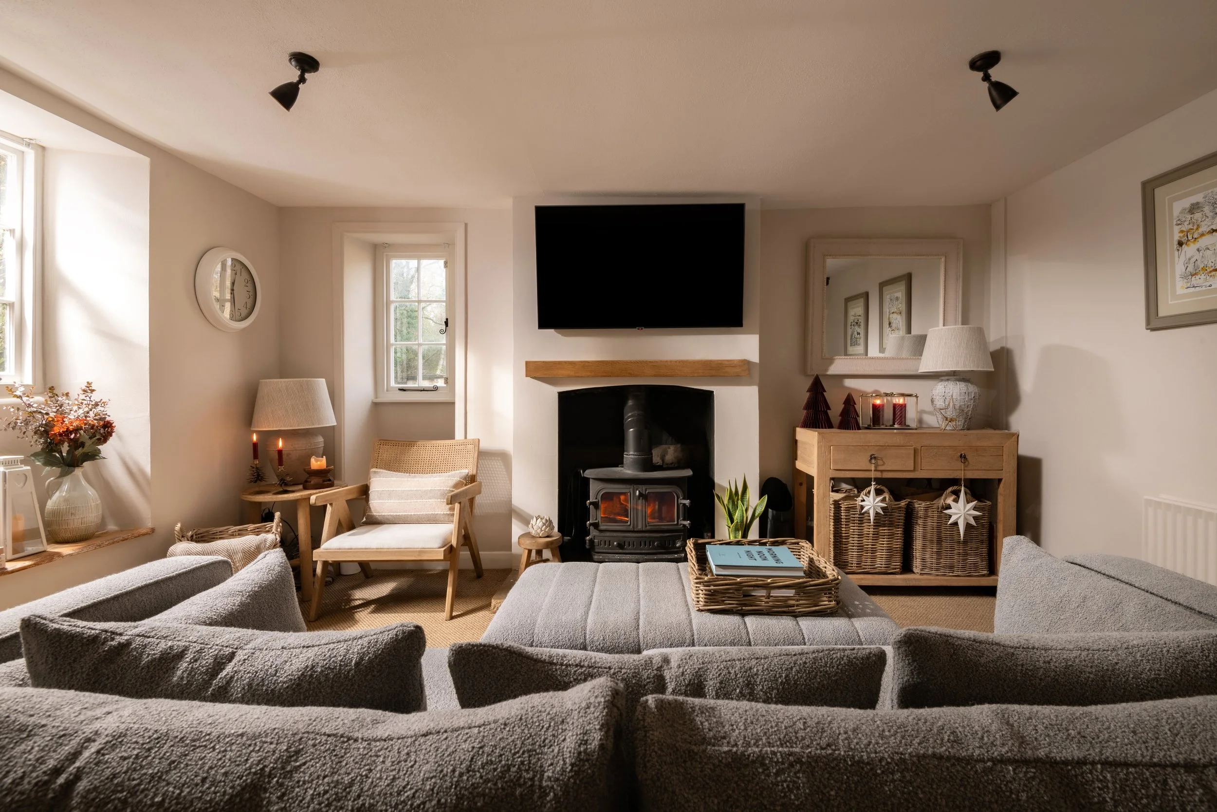 Living room with a fireplace, TV mounted above, beige armchair by the window, gray sofa, wooden side table with candles, framed artwork, decorative mirror, basket, potted plant, and books.