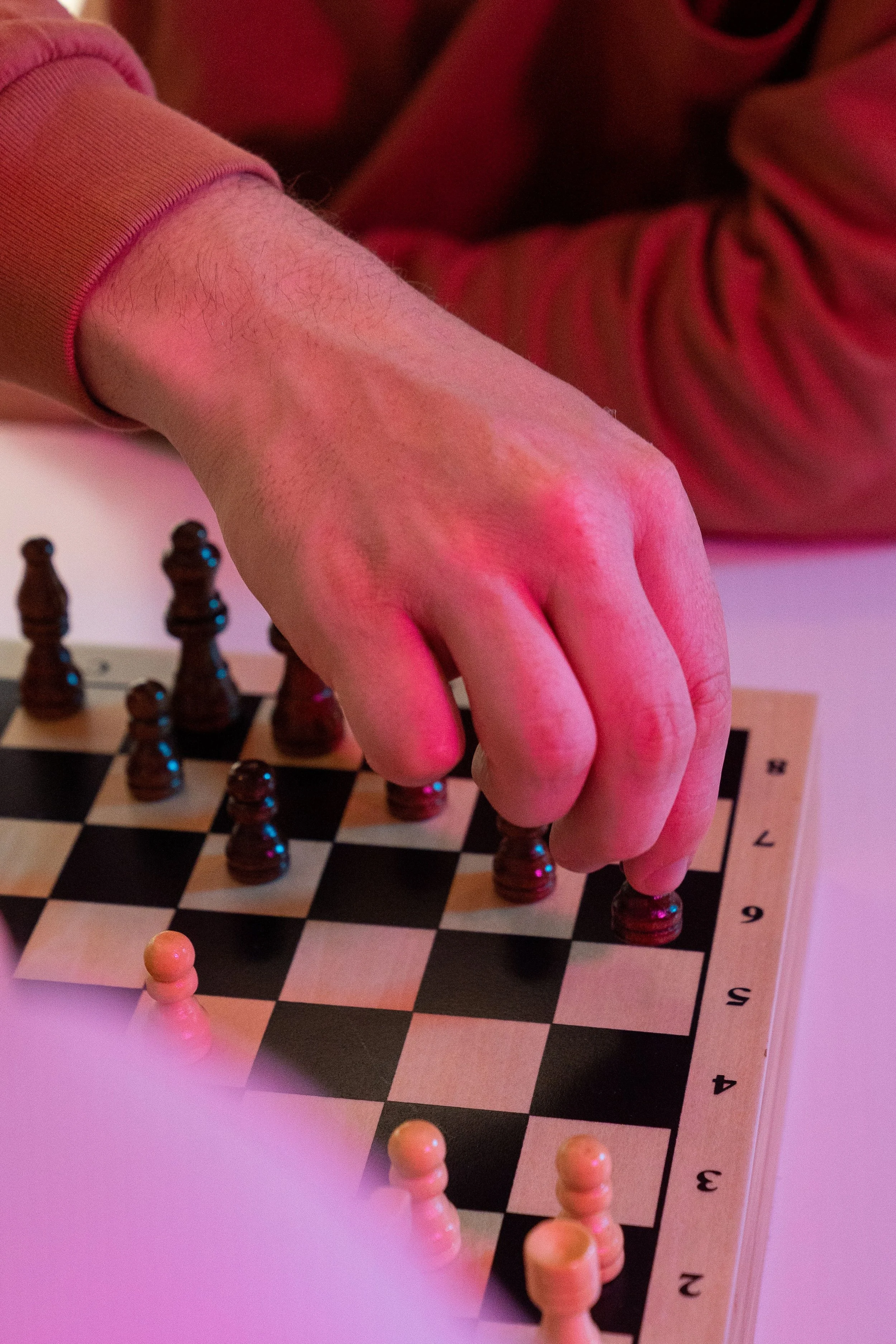 A person’s hand moving a black chess piece on a chessboard with numbered rows and columns, illuminated by pinkish lighting.