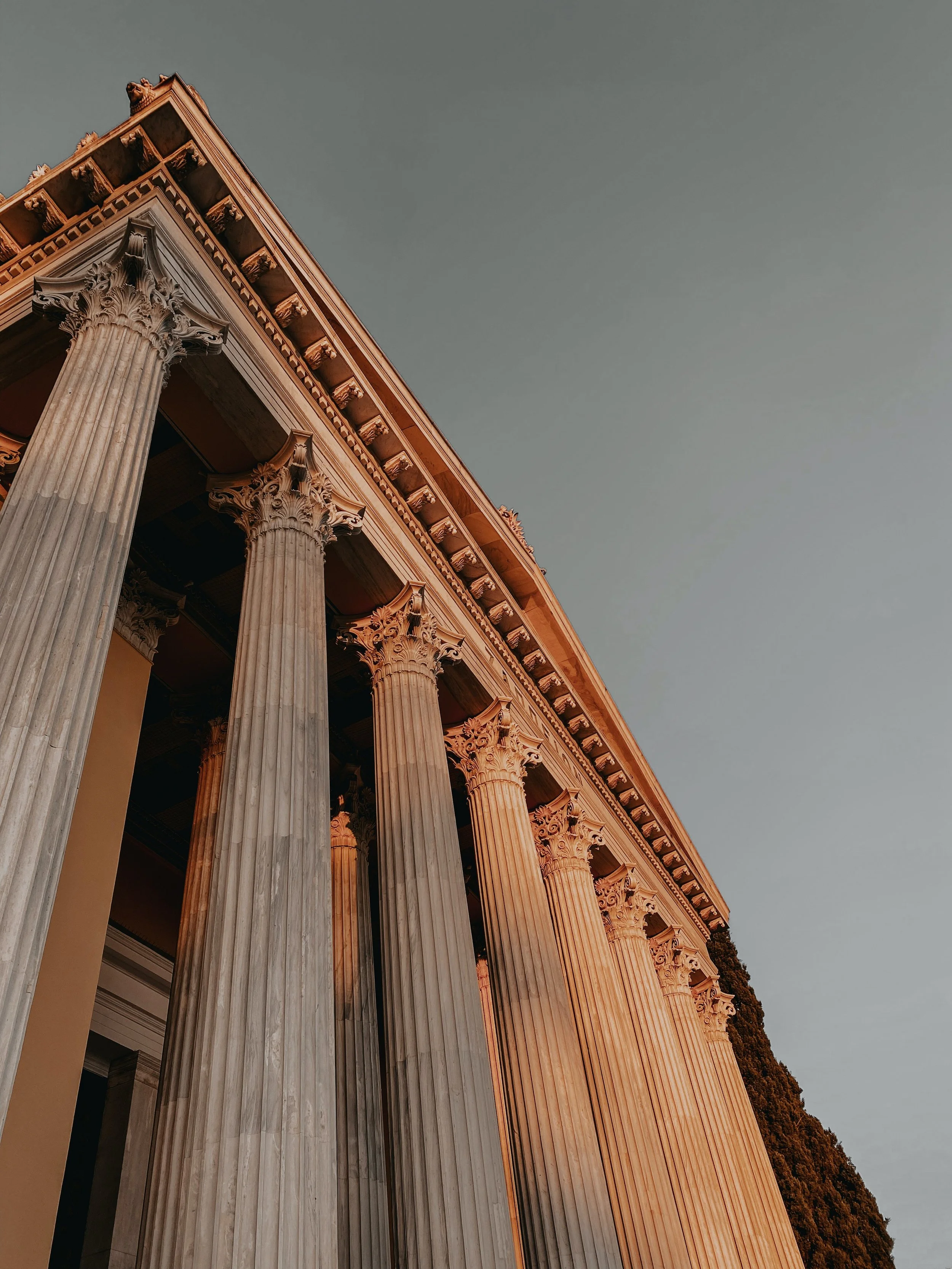 A classical Greek temple with tall fluted columns and ornate capitals, part of a larger structure, with a clear sky in the background.