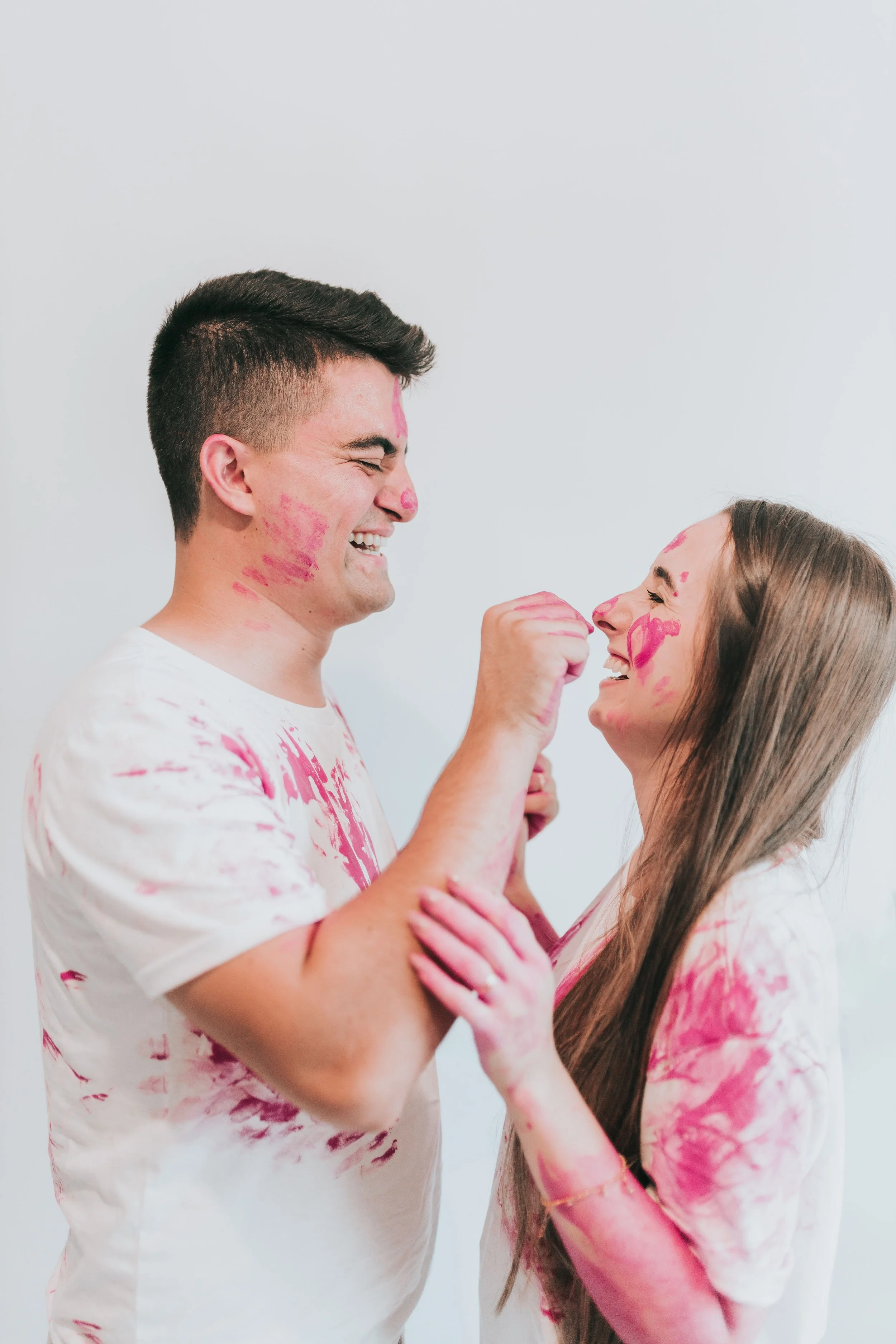 A young man and woman with painted faces and matching shirts, smiling and playfully touching each other's faces against a plain white background.