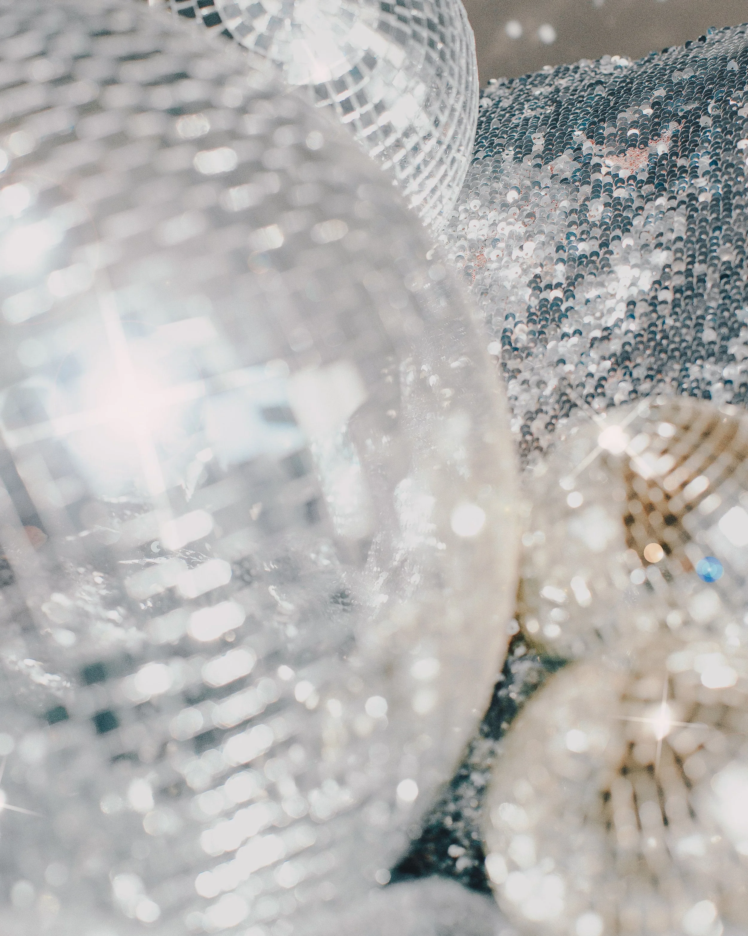 Close-up of a glittering disco ball reflecting light, with a background of many small shiny sequins or beads creating a shimmering surface.