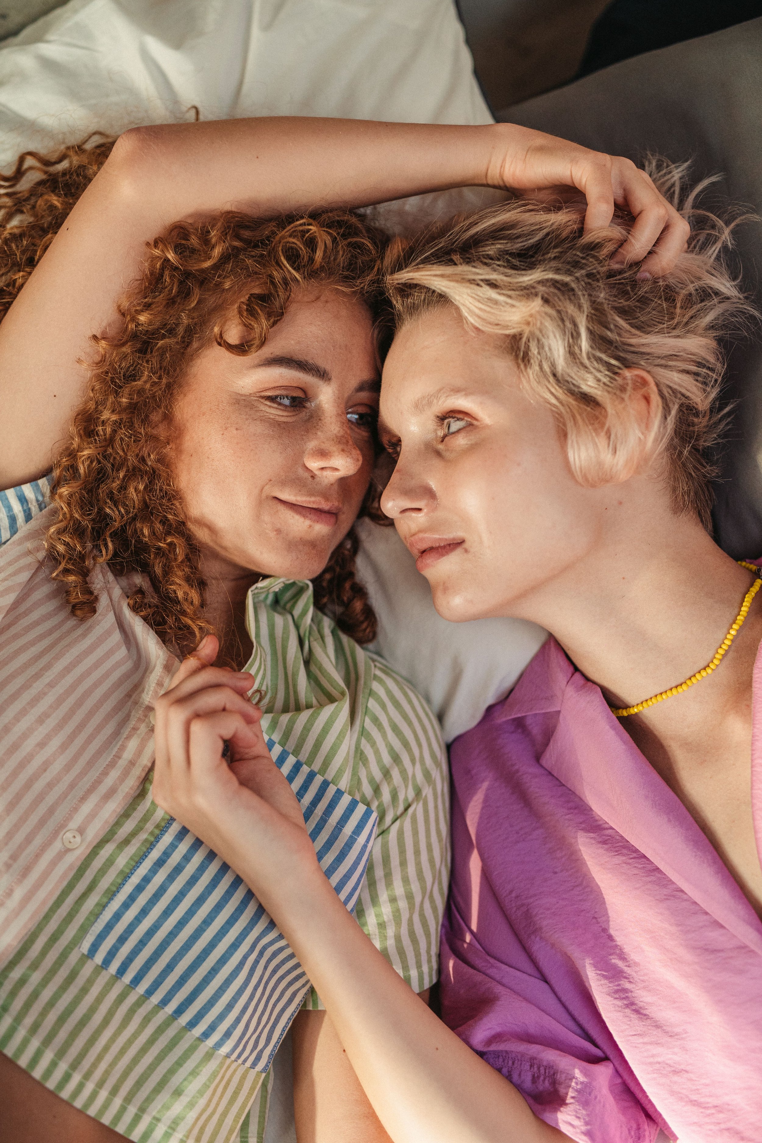 Two women with curly and short blonde hair lying close together, looking into each other's eyes, touching hands, in bed, wearing colorful pajamas.