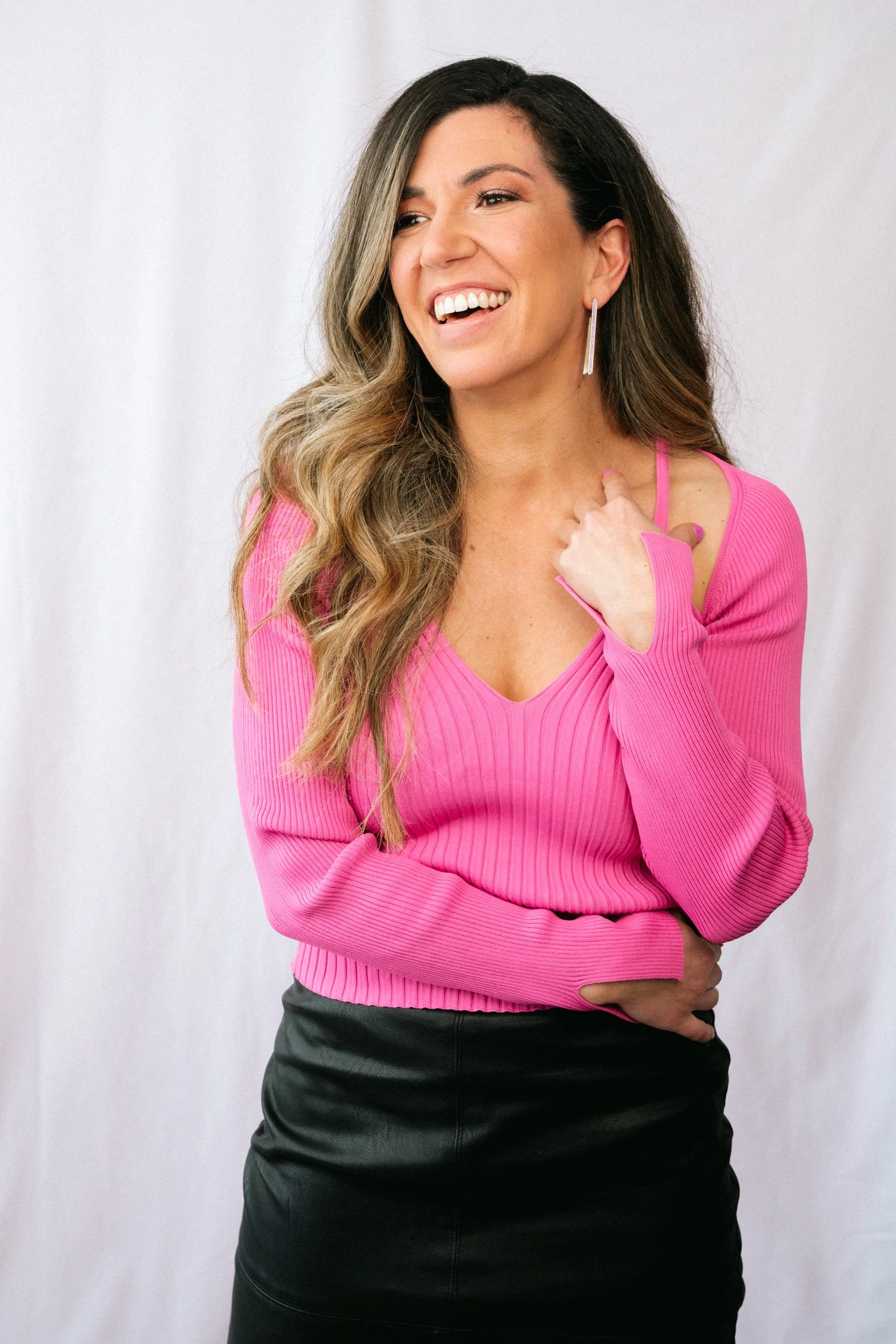 A woman wearing a pink long-sleeve top and a black skirt poses against a plain white wall, smiling, with one hand on her chest and the other across her body.