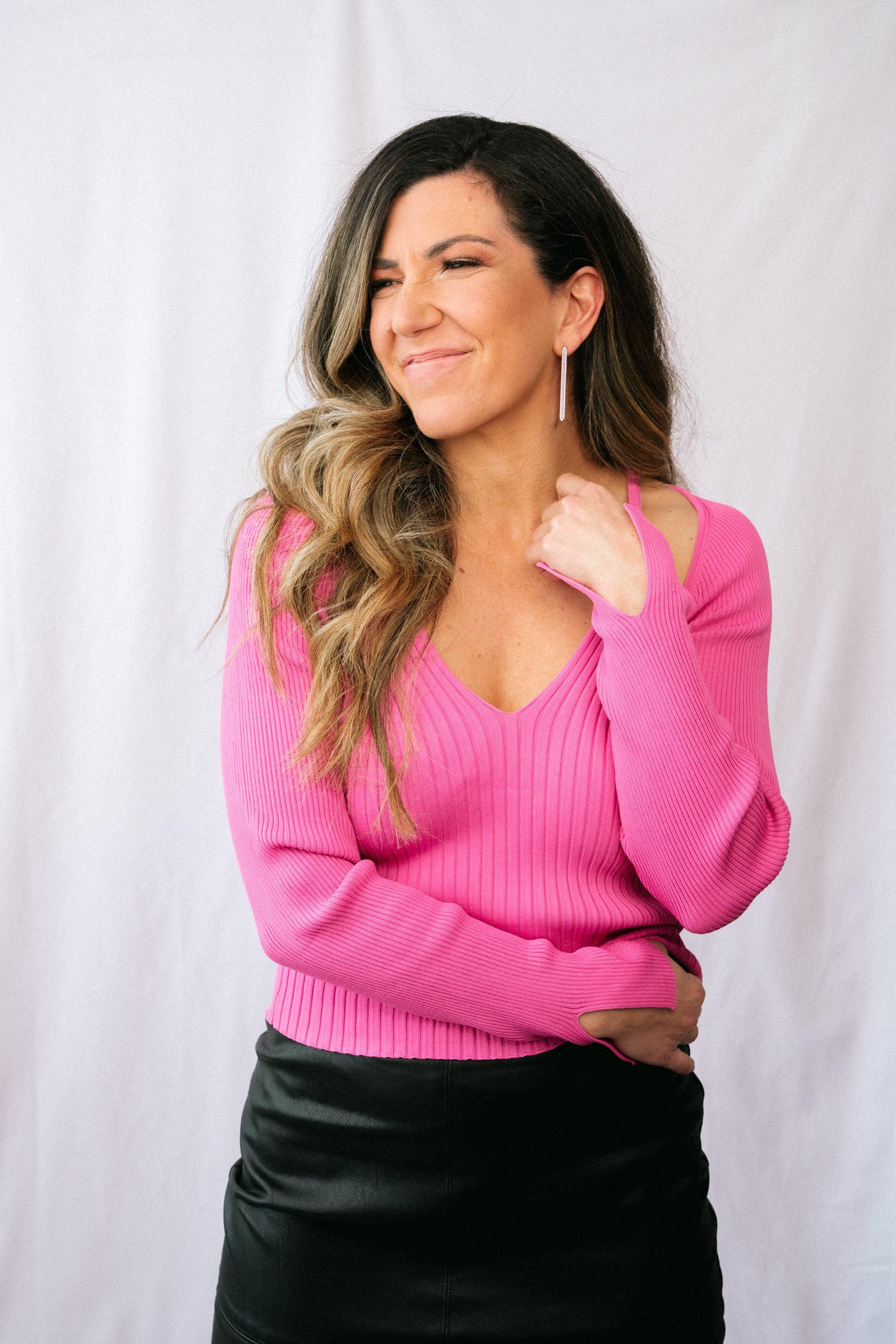 A woman with long, wavy hair wearing a pink ribbed sweater, silver dangle earrings, and black skirt, standing against a plain, light-colored background with a smile and a playful pose.