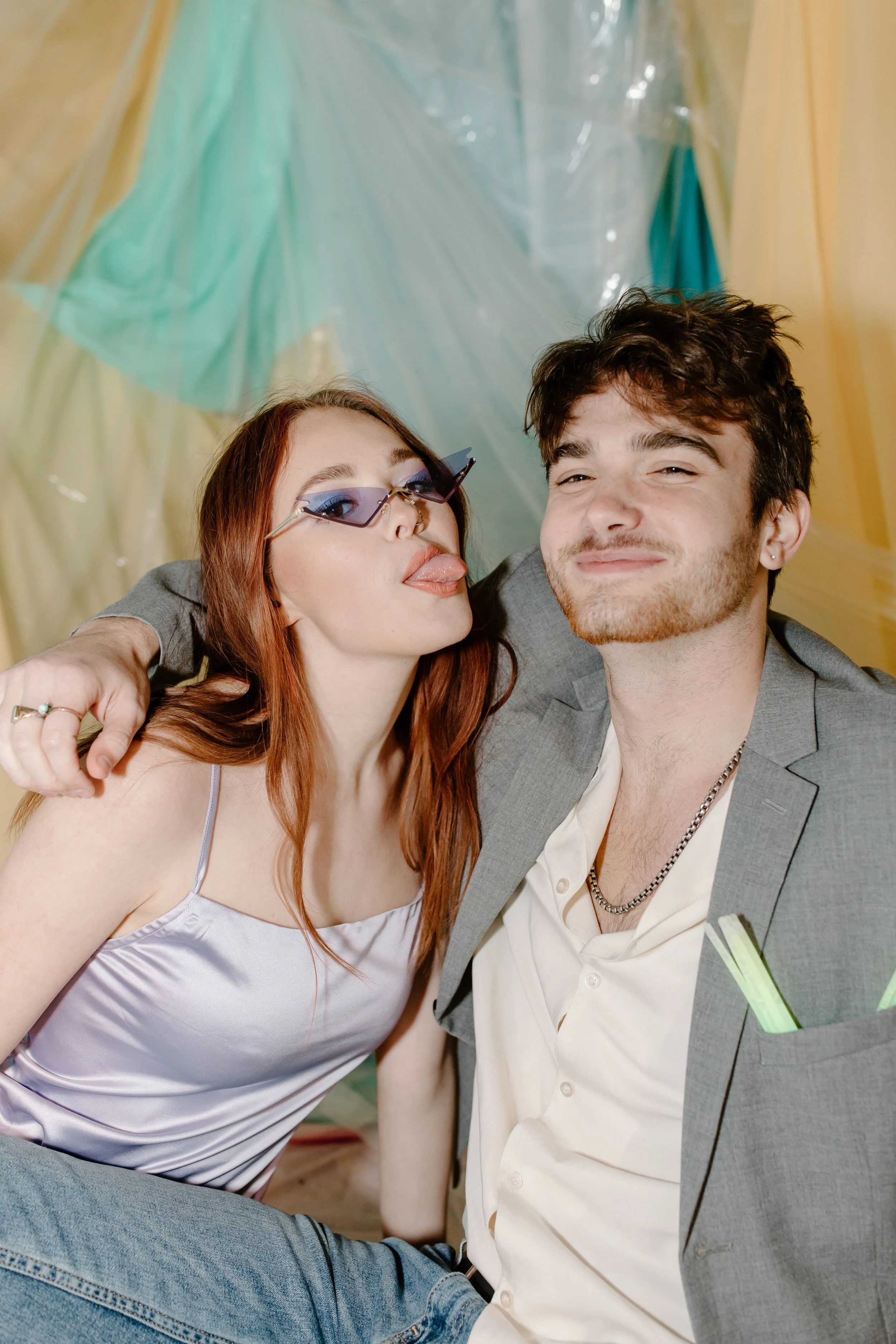 A young woman with red hair and wearing sunglasses sticks her tongue out while taking a selfie with a young man with dark hair. The woman is dressed in a light-colored satin top, and the man is dressed in a gray blazer with a white shirt and layered necklaces. They are smiling and appear to be happy, with a colorful background behind them.