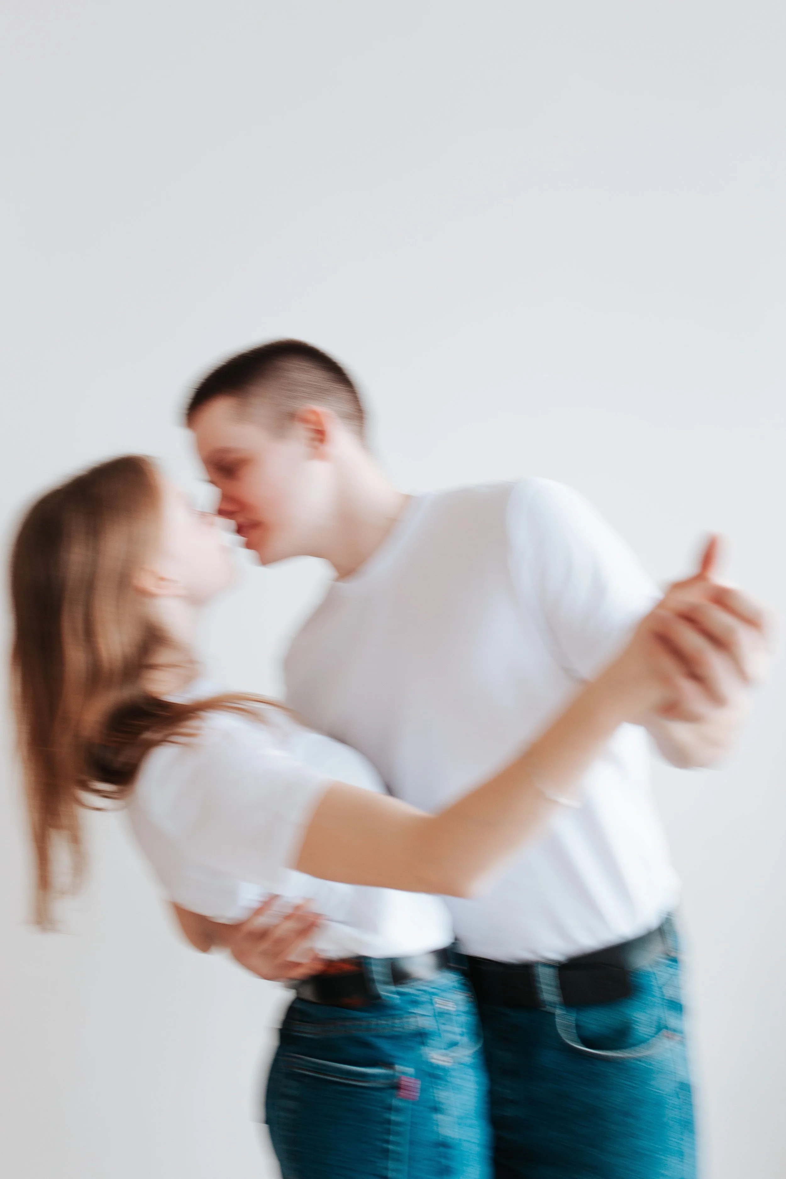 A young man and woman are close together, engaged in a dance or intimate moment, with their eyes closed and faces near each other, against a plain white background.