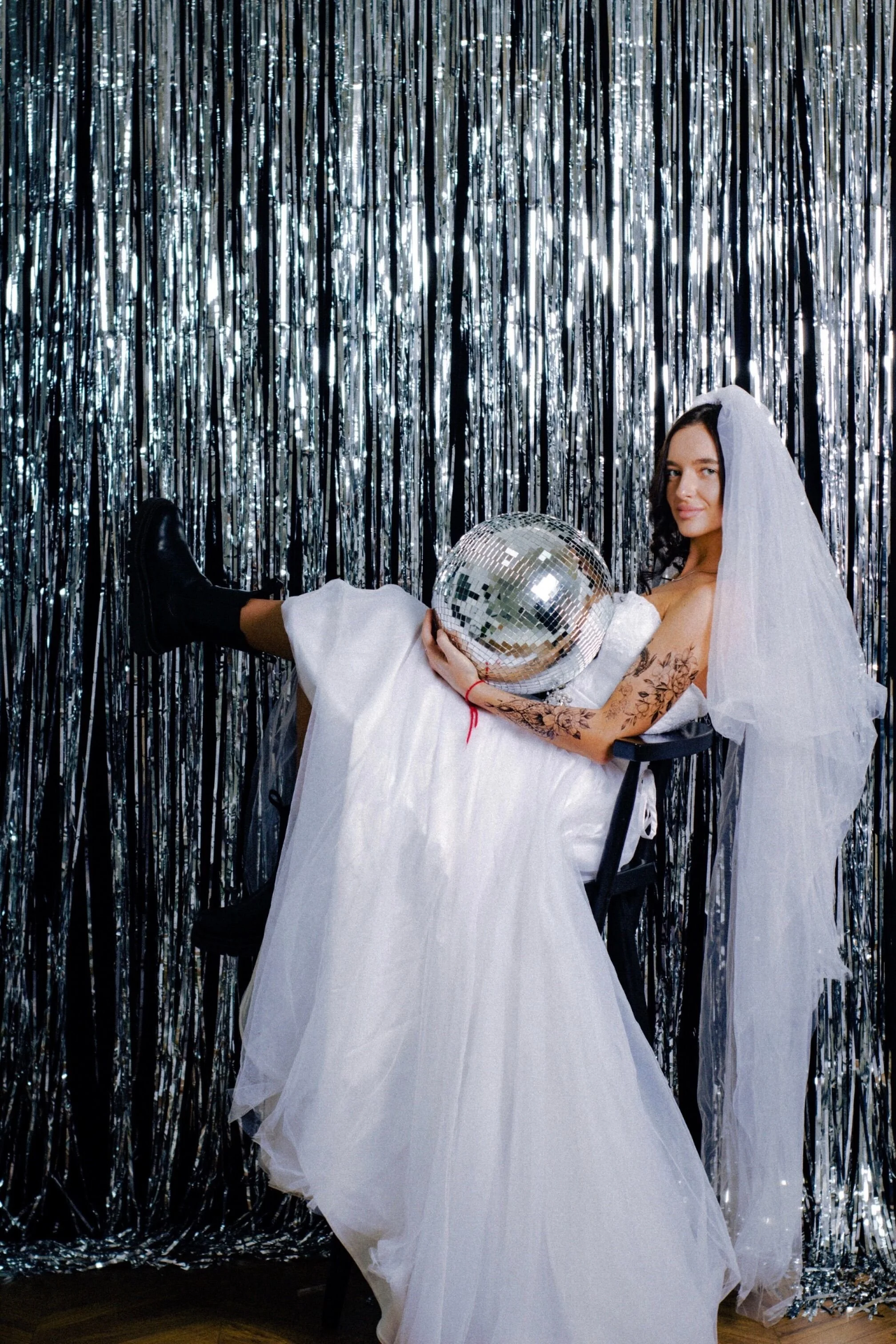 A woman in a wedding dress and veil lying on a chair, holding a disco ball, with a silver tinsel curtain backdrop.