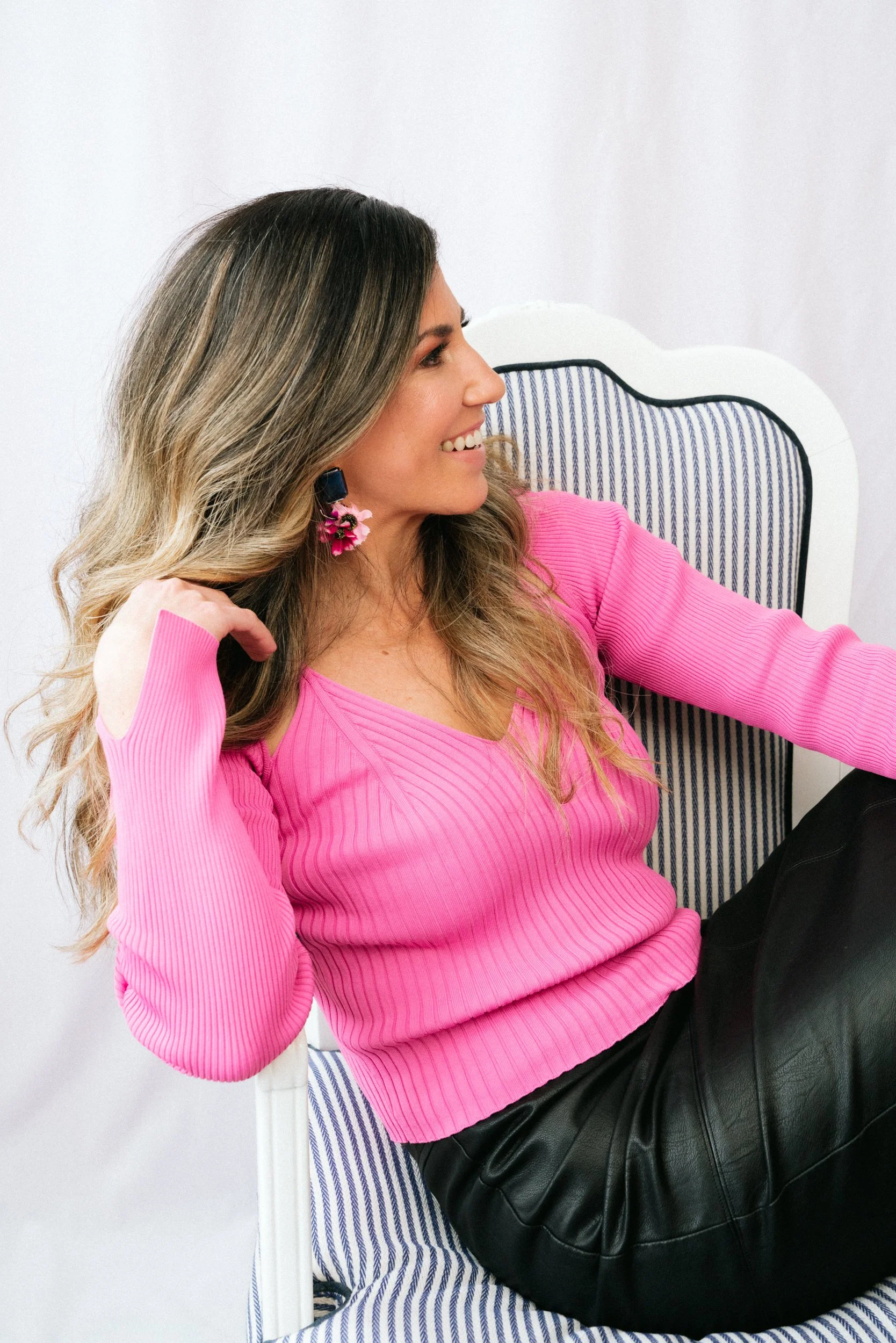 A woman with long, wavy hair sitting on a chair with a striped backrest, wearing a bright pink long-sleeve top and black leather skirt, smiling and looking to her left.