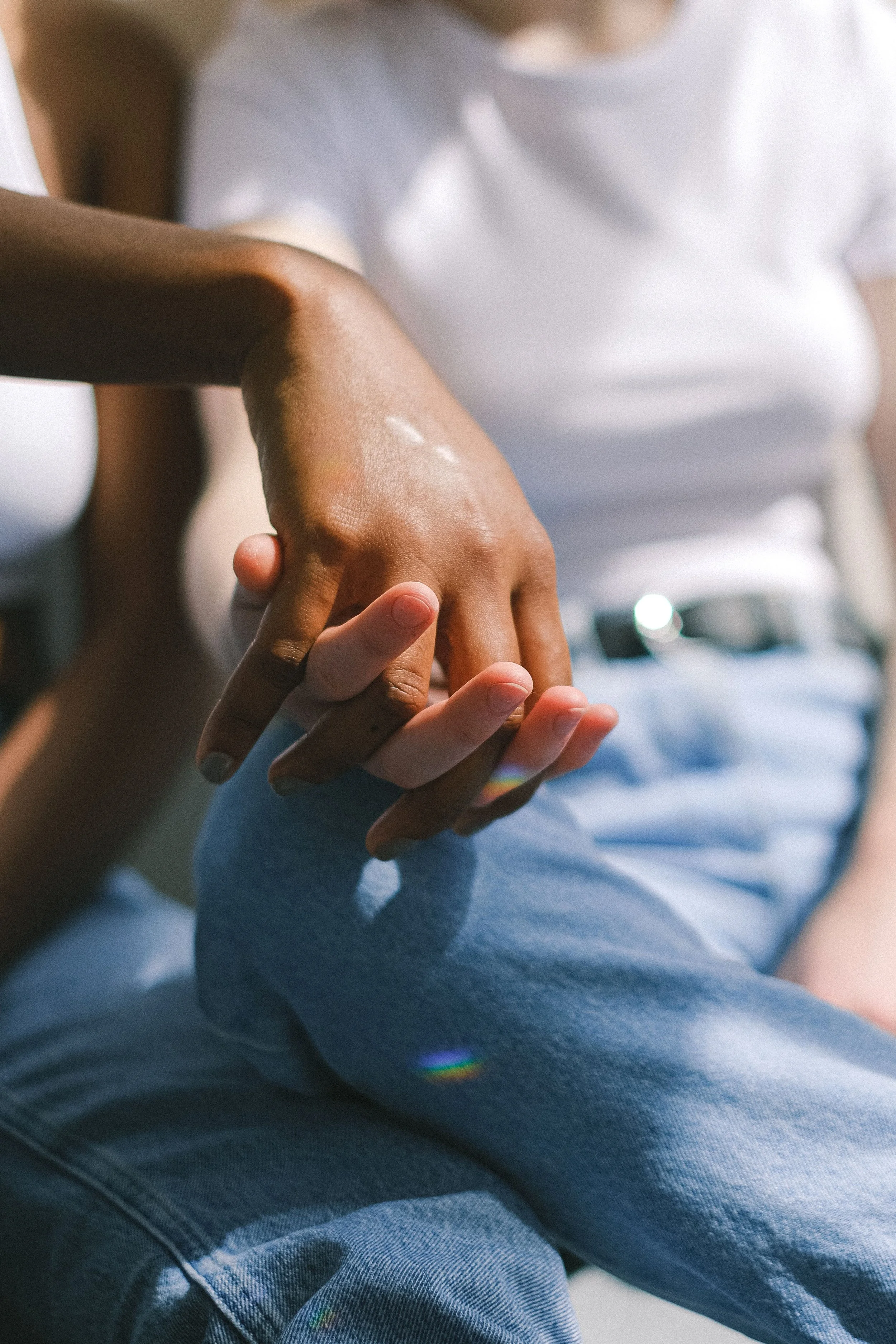 Close-up of two people holding hands, one with dark skin and the other with light skin, sitting outdoors. The person with dark skin is wearing a white shirt, and the person with light skin is wearing blue jeans. Sunlight creates a small rainbow reflection on the leg of the person with light skin.