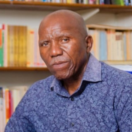 A mature man with a serious expression, wearing a blue patterned shirt, sitting in front of a bookshelf filled with books.