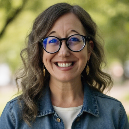 Smiling woman with wavy brown hair, blue glasses, gold earrings, wearing a denim jacket outdoors with blurred green foliage in the background.