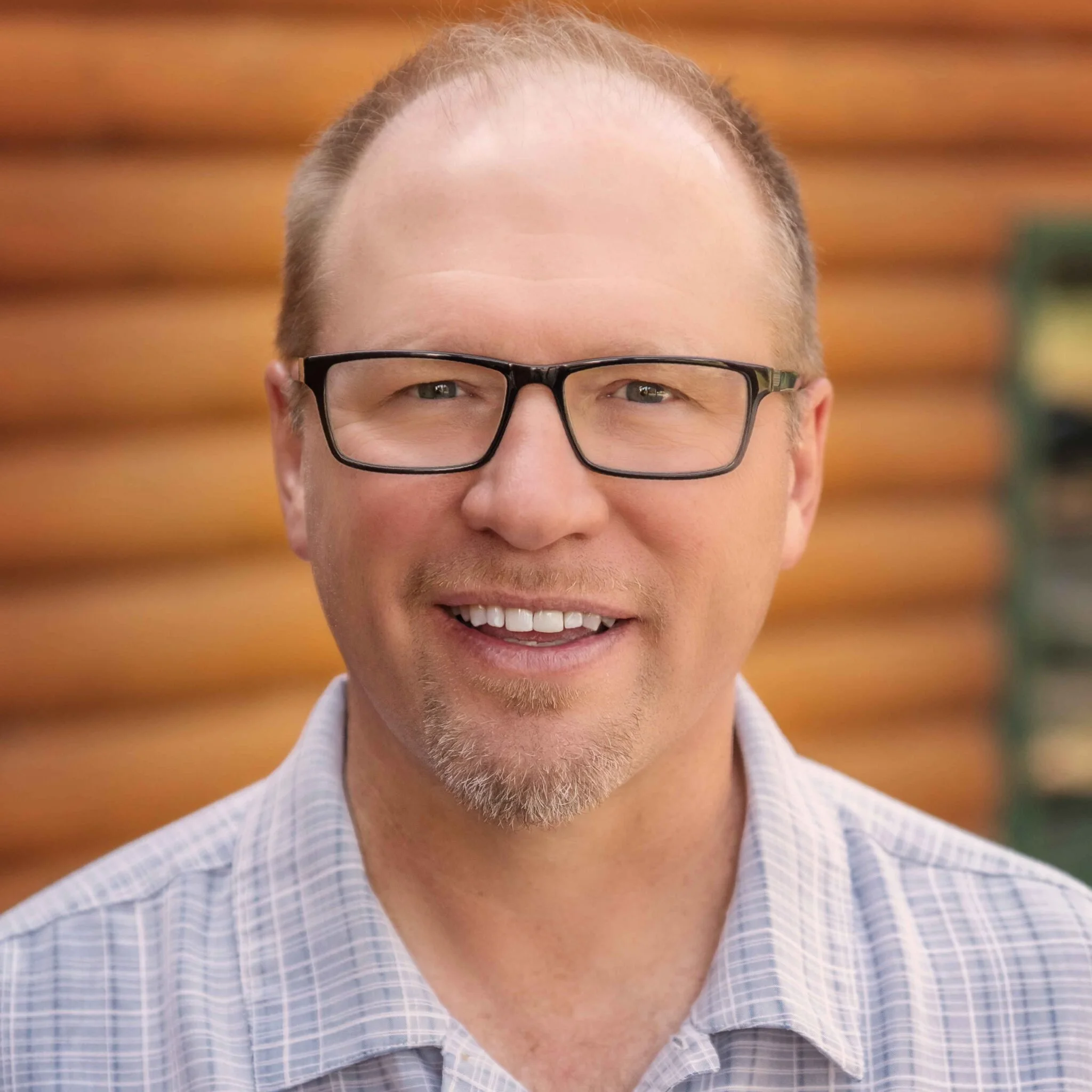 Close-up of a smiling man with glasses, a bald head, and a light beard, wearing a blue and white checkered shirt, with a blurred wooden background.