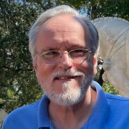 A smiling man with glasses and a beard, wearing a blue shirt outdoors with trees and a sculpture in the background.