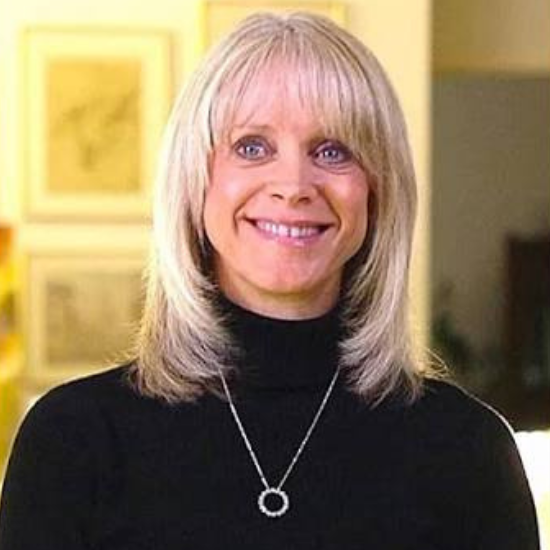A woman with blonde hair, wearing a black turtleneck and a circular pendant necklace, smiling indoors.