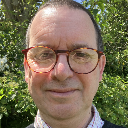Close-up of a man with glasses smiling outdoors with green foliage in the background.